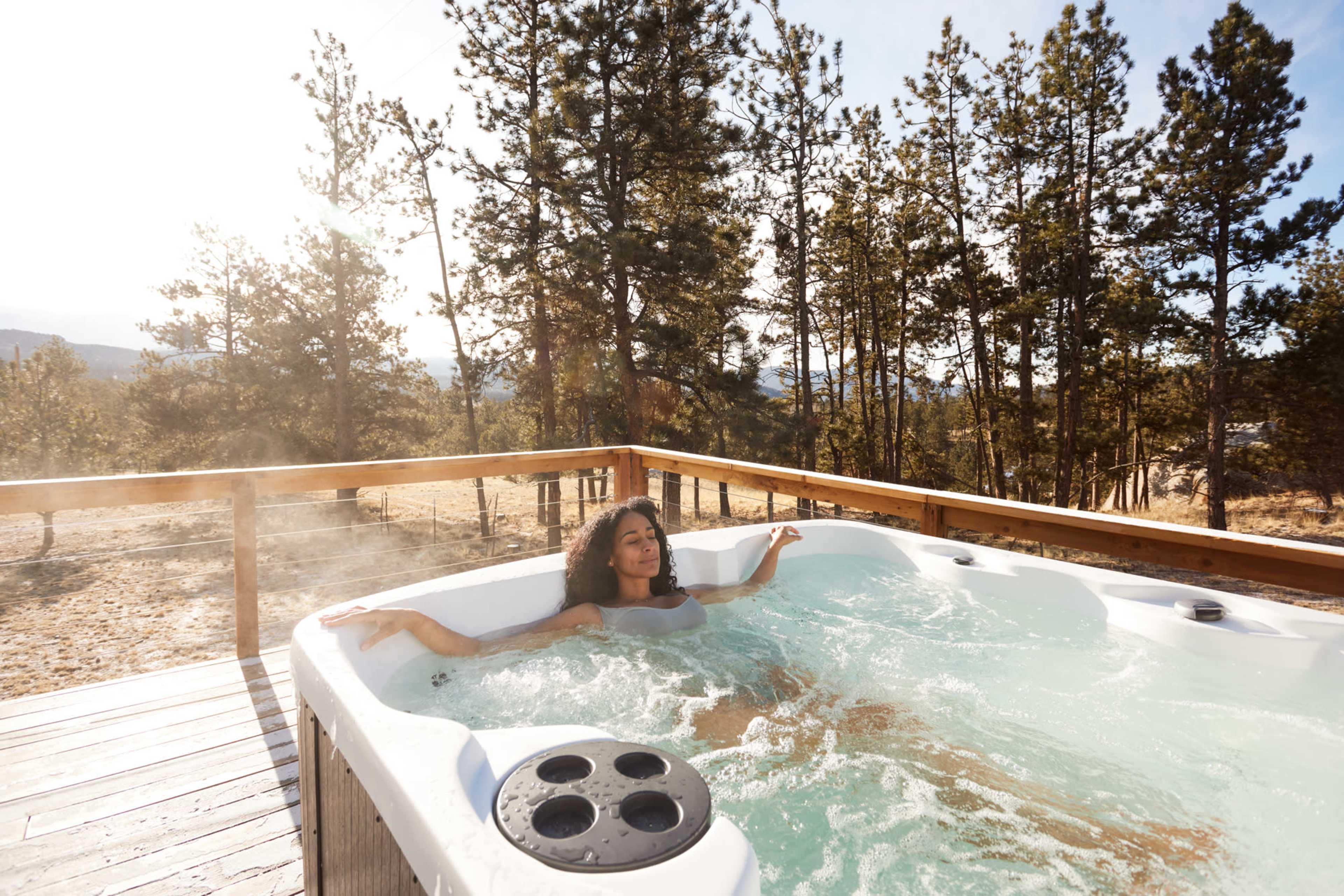 A woman relaxes in a hot tub on a wooden deck surrounded by trees and mountains.
