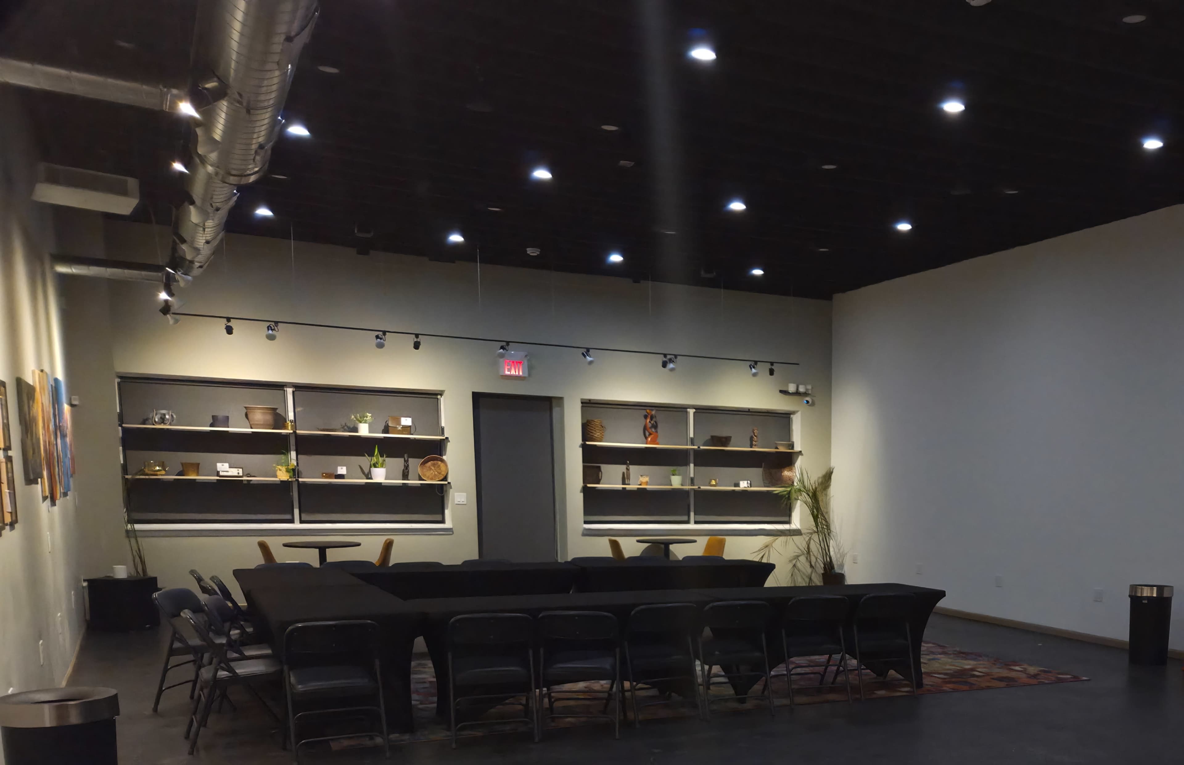 The image shows a well-lit, spacious room with a circular arrangement of chairs around a dark table, flanked by shelves displaying various decorative items.