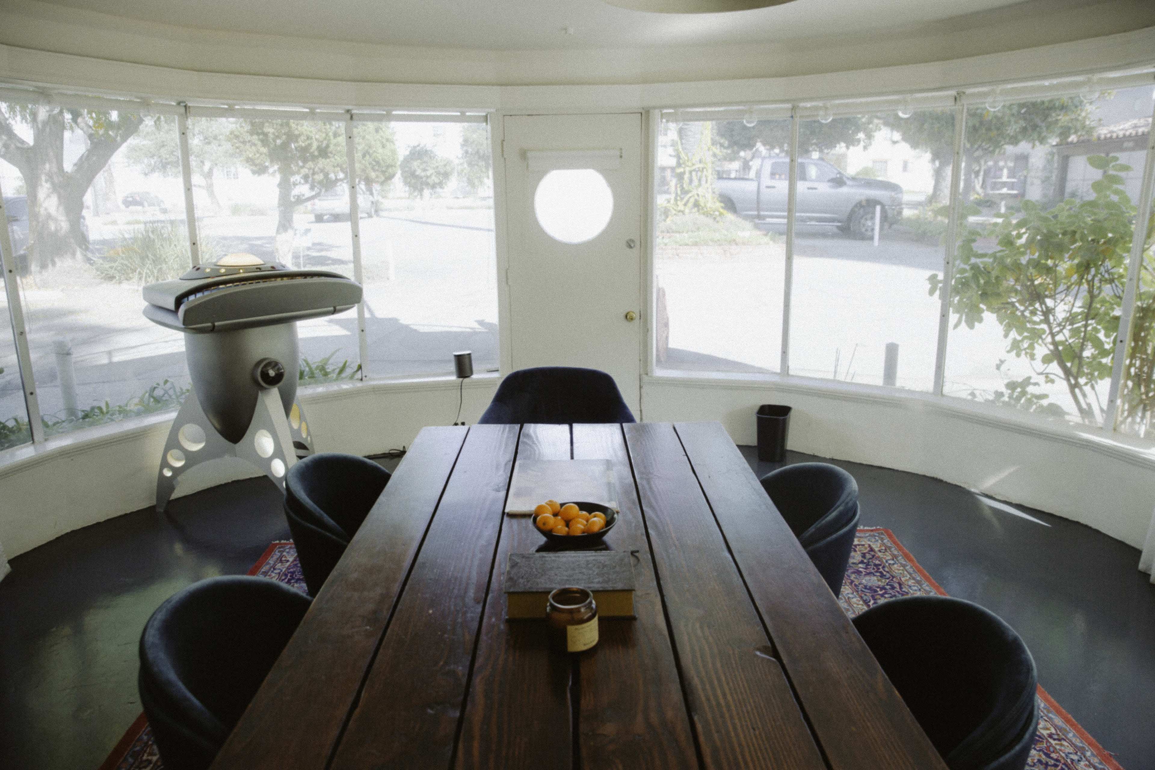 A circular dining area features a large wooden table surrounded by dark chairs, with a round window and a visible outdoor scene.
