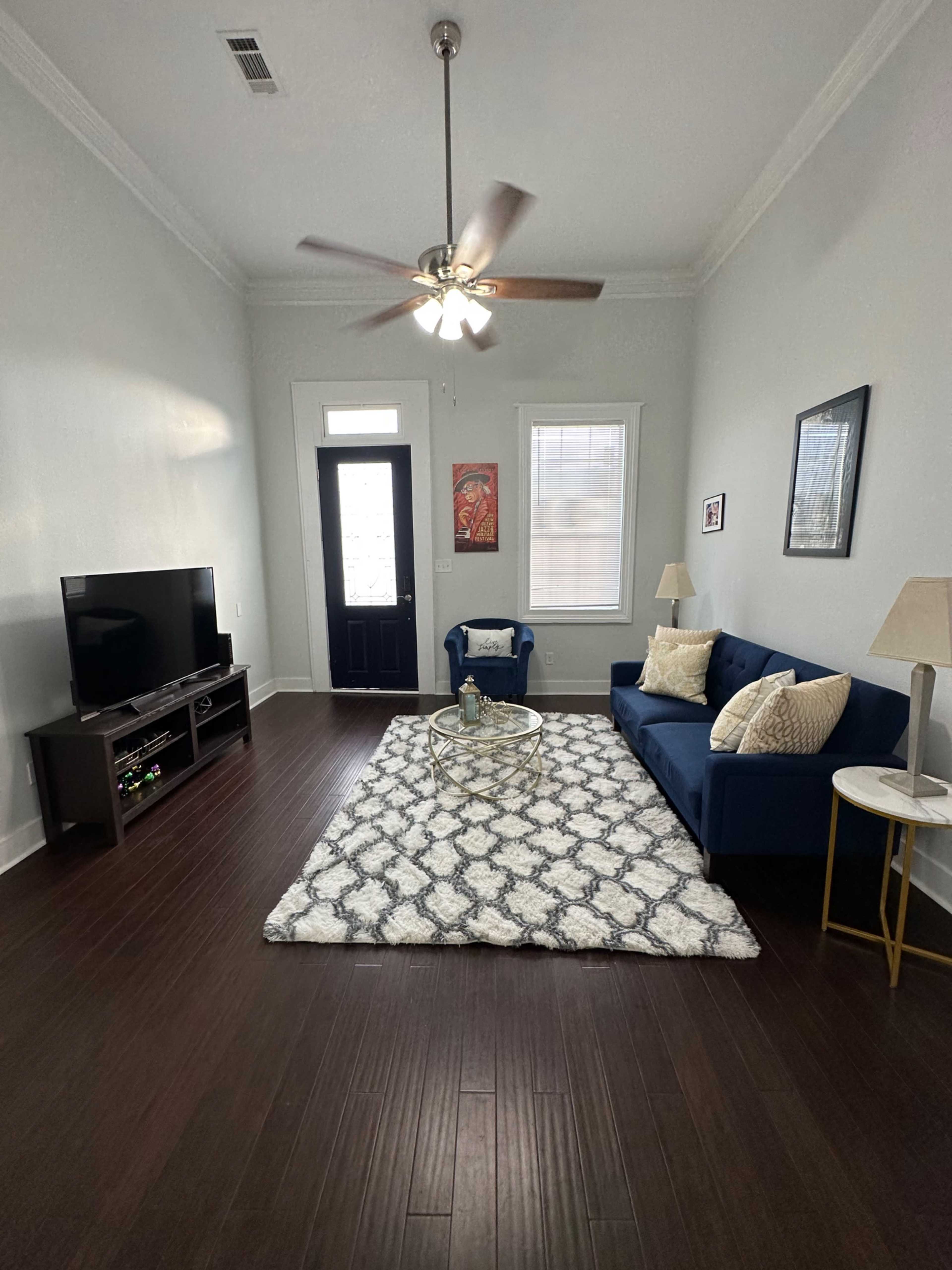The living room features a blue sofa, a round coffee table on a patterned rug, a television unit, and a front door with windows.