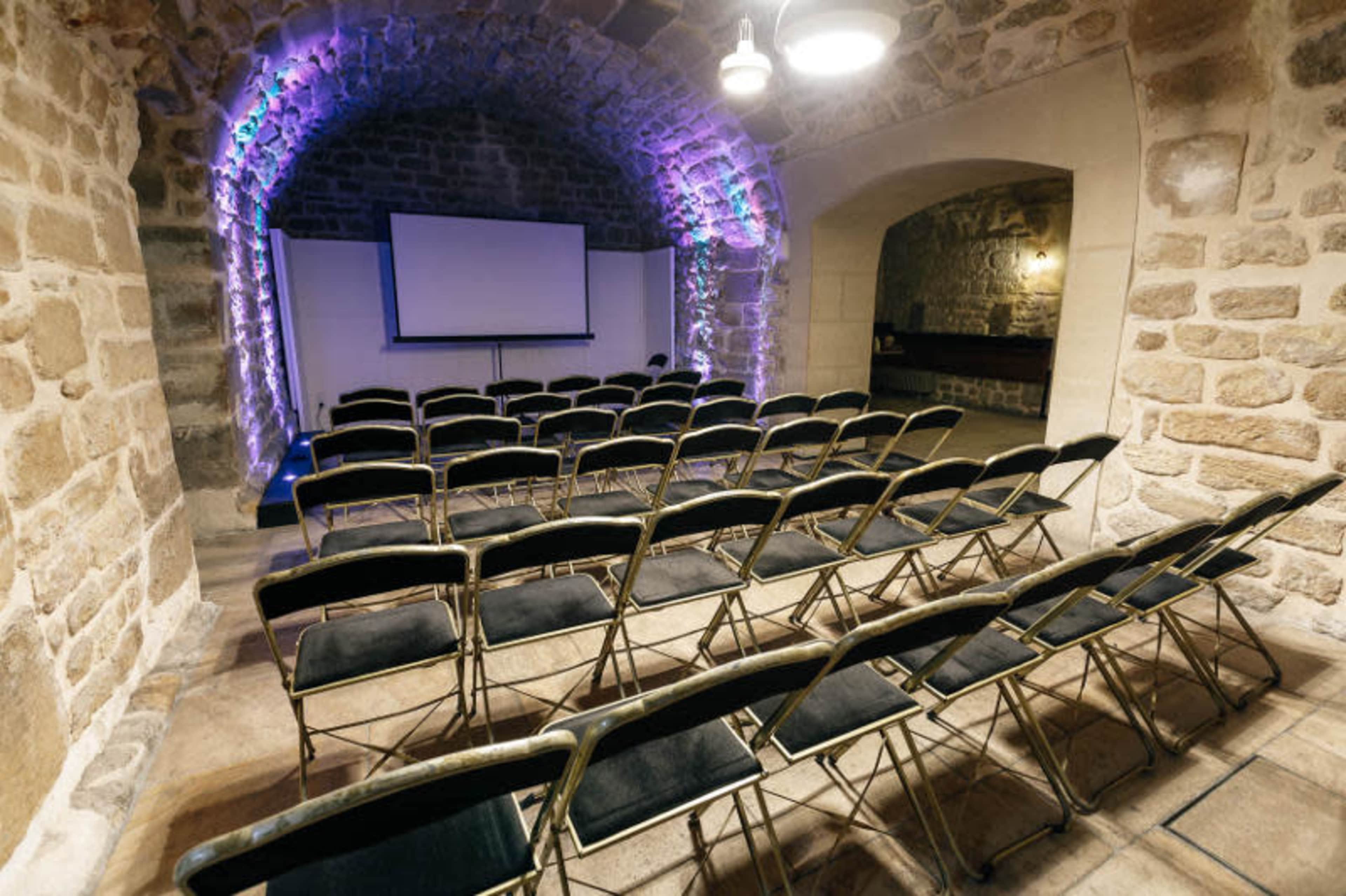 A stone-walled meeting room features rows of black folding chairs facing a projection screen.