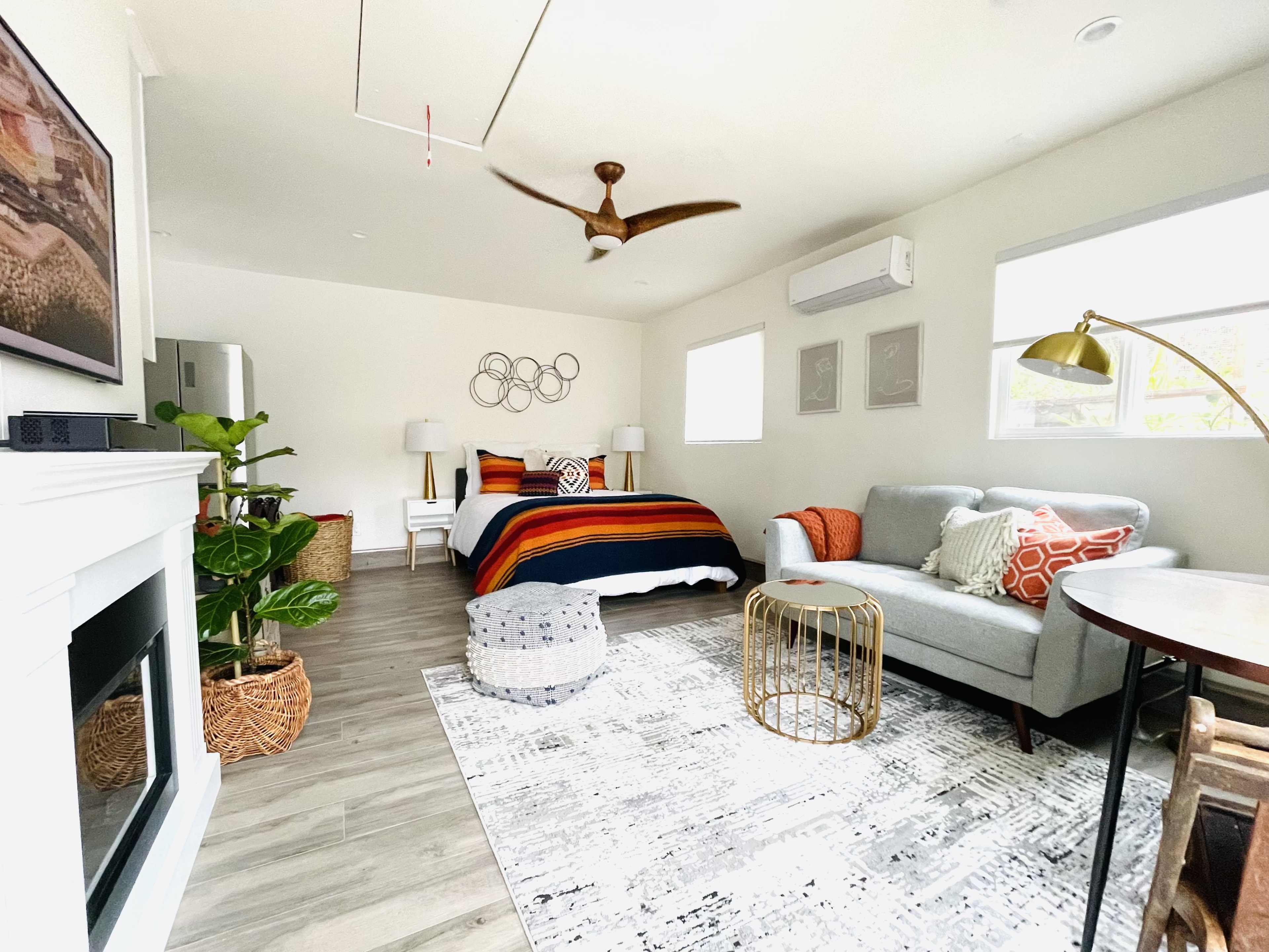 The image shows a neatly arranged bedroom with a bed covered in a striped blanket, a green plant in the corner, a light gray sofa, and decorative pillows, set against light-colored walls and wooden flooring.