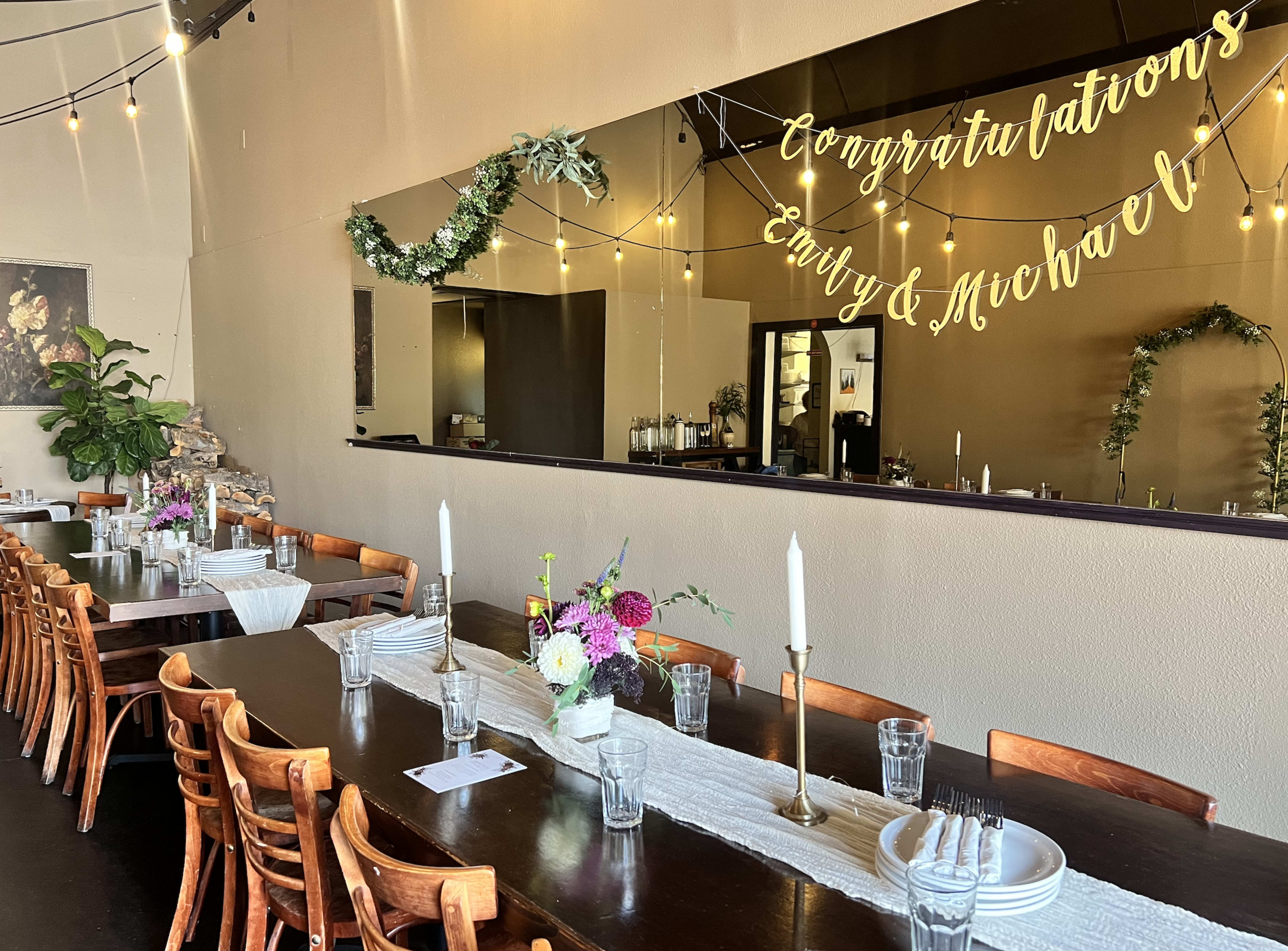 The image shows a dining setup in a restaurant with a long table adorned with floral arrangements, candles, and decorations celebrating "Emmy & Michael."