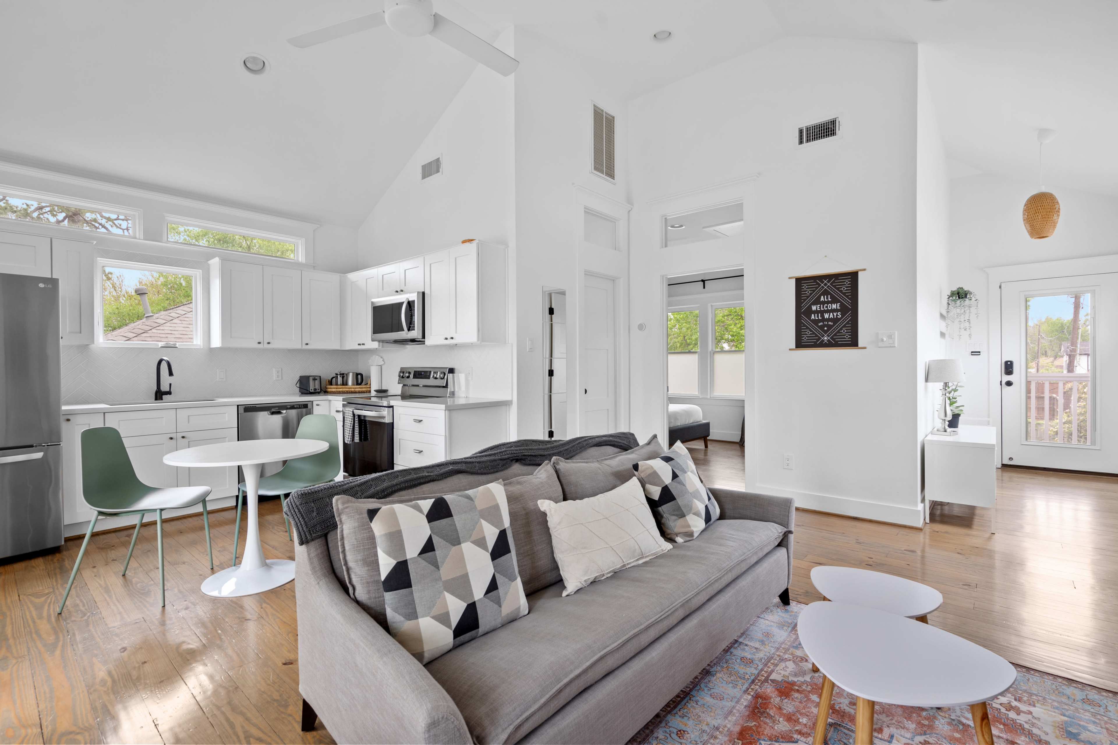 The image shows a modern, open-concept living area with a couch, two small tables, a dining space, and a kitchen featuring white cabinetry and stainless steel appliances.