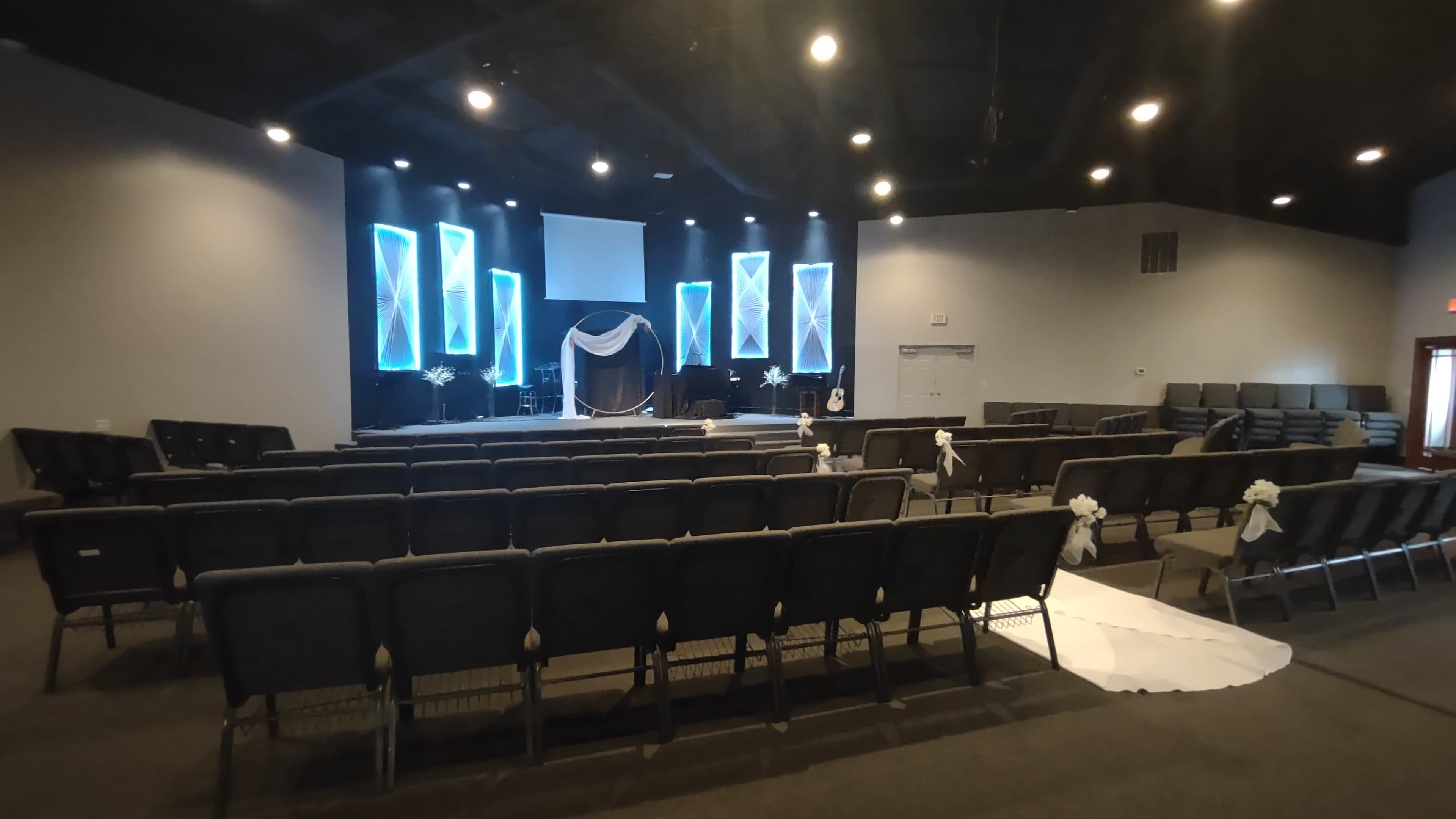 The interior of a theater-style venue features rows of gray chairs facing a stage adorned with lit panels and decorative drapery.
