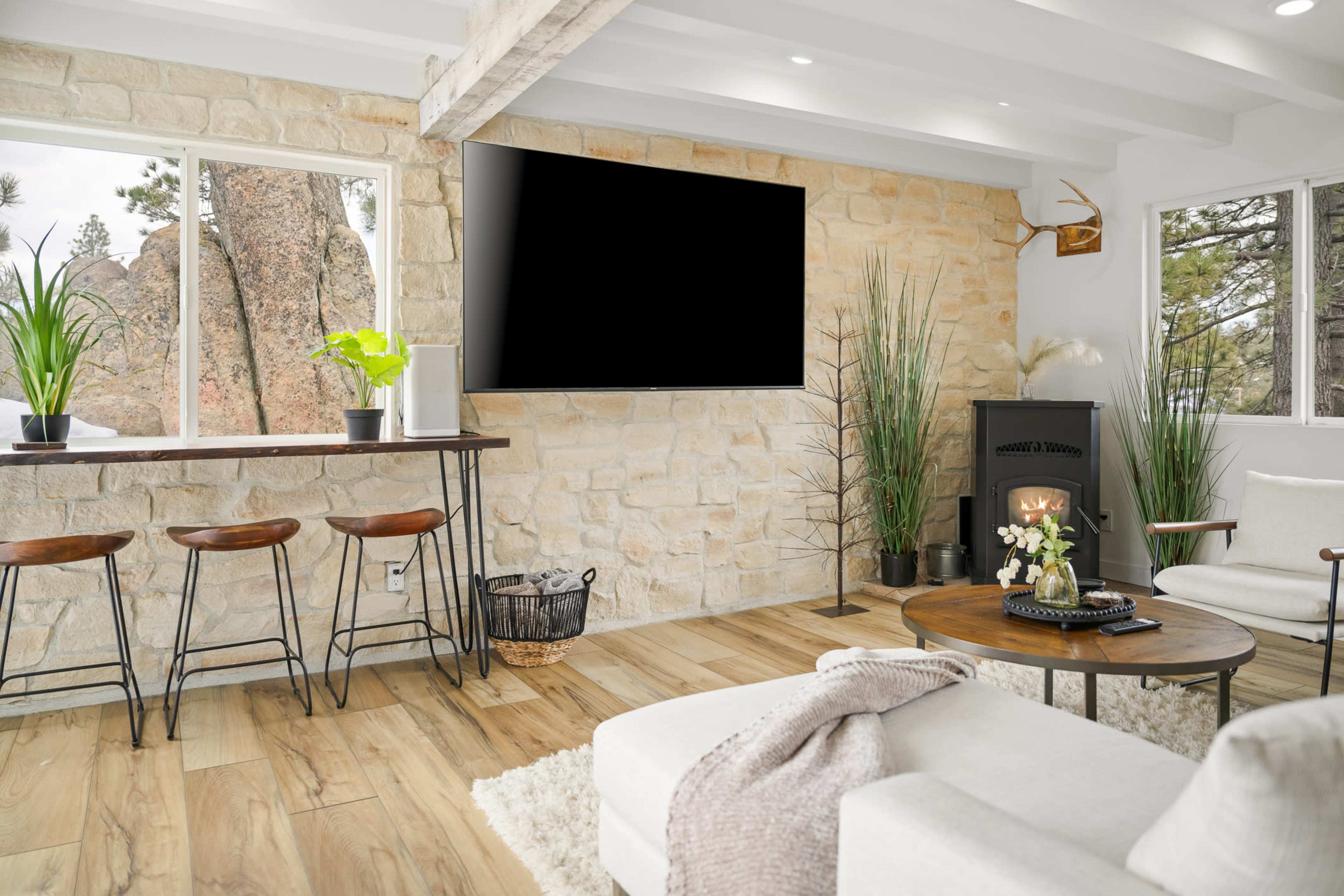 The image shows a modern living room with a stone wall, a flat-screen TV, a wooden bar with stools, a round table, and a cozy seating area near a small stove.