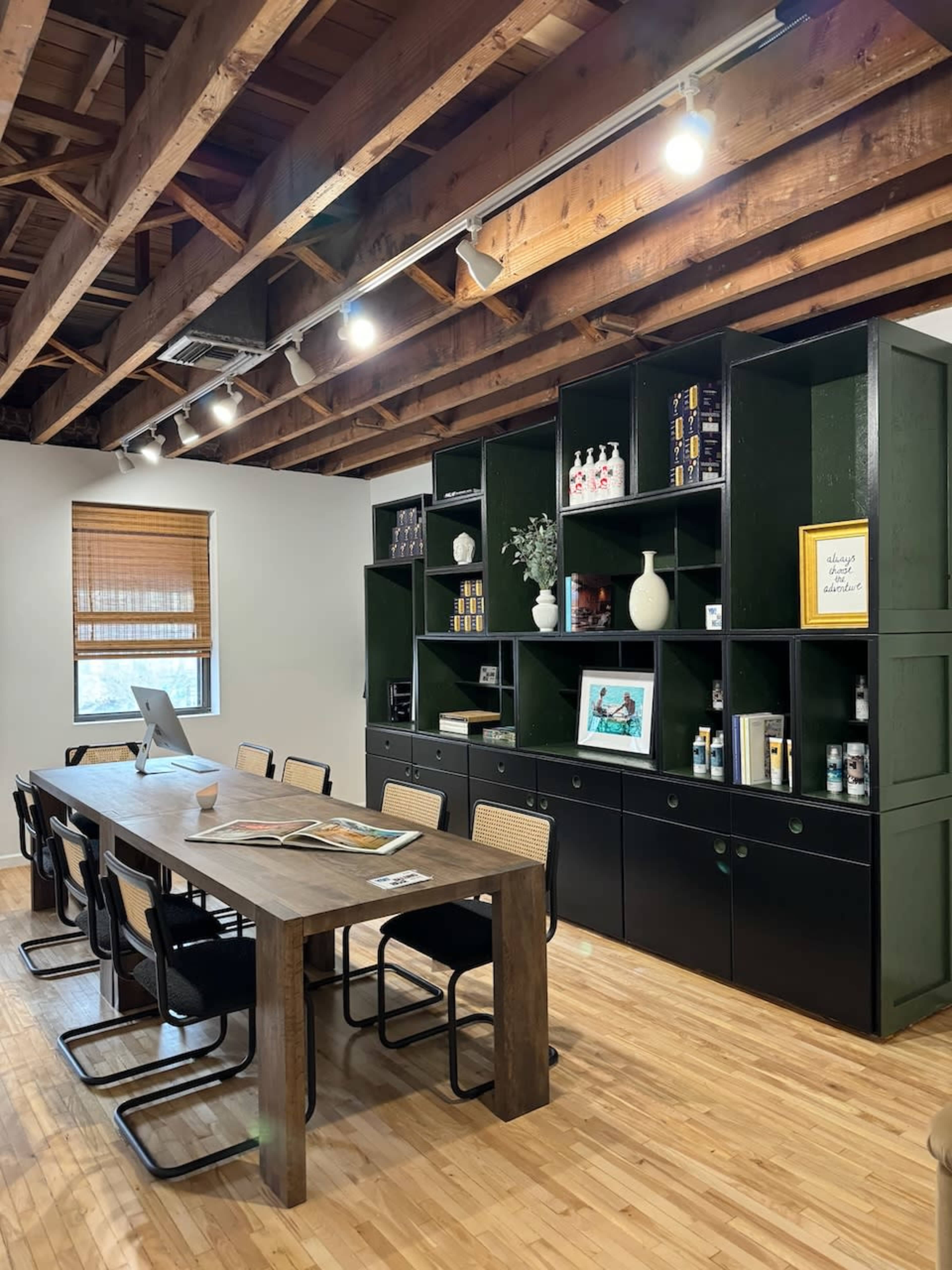 The image depicts a modern office space featuring a long wooden table surrounded by black chairs, with a green shelving unit displaying various items in the background.