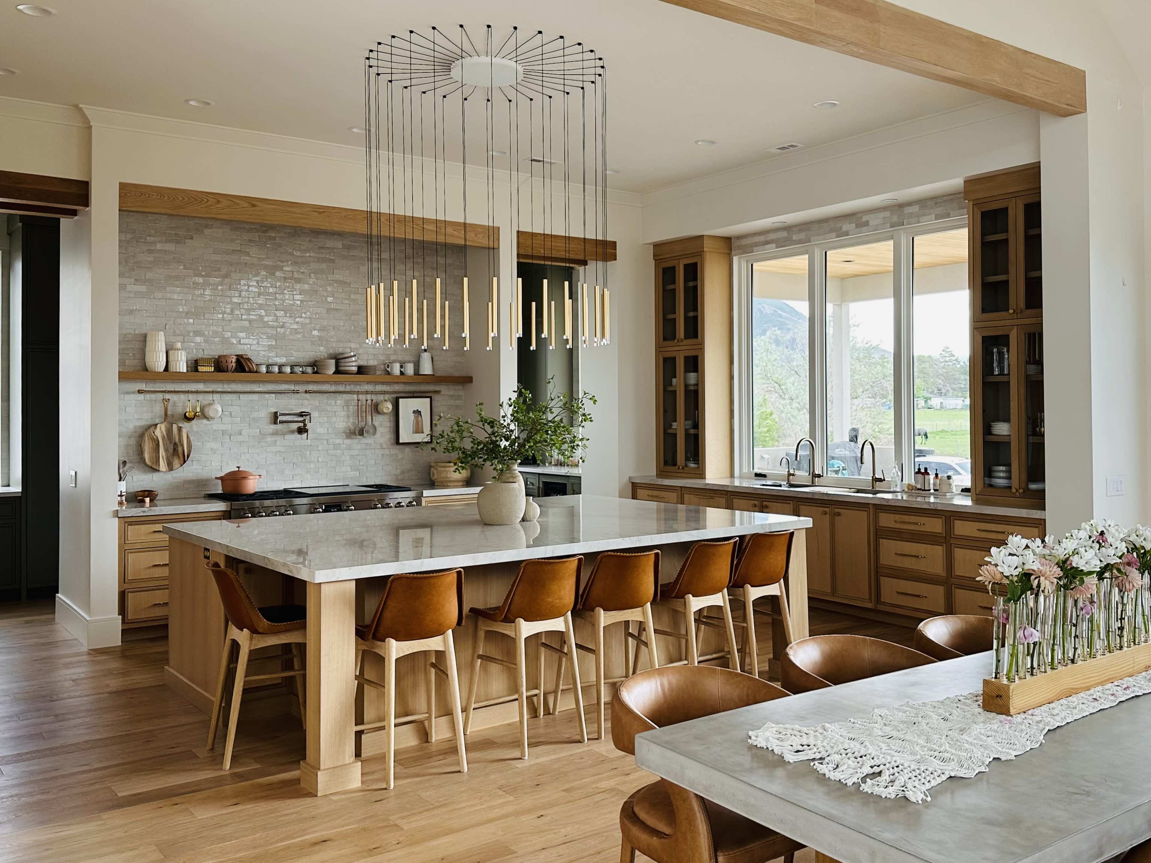 The image shows a modern kitchen with a large island, wooden cabinetry, and a spacious dining area featuring a decorative table and floral arrangements.