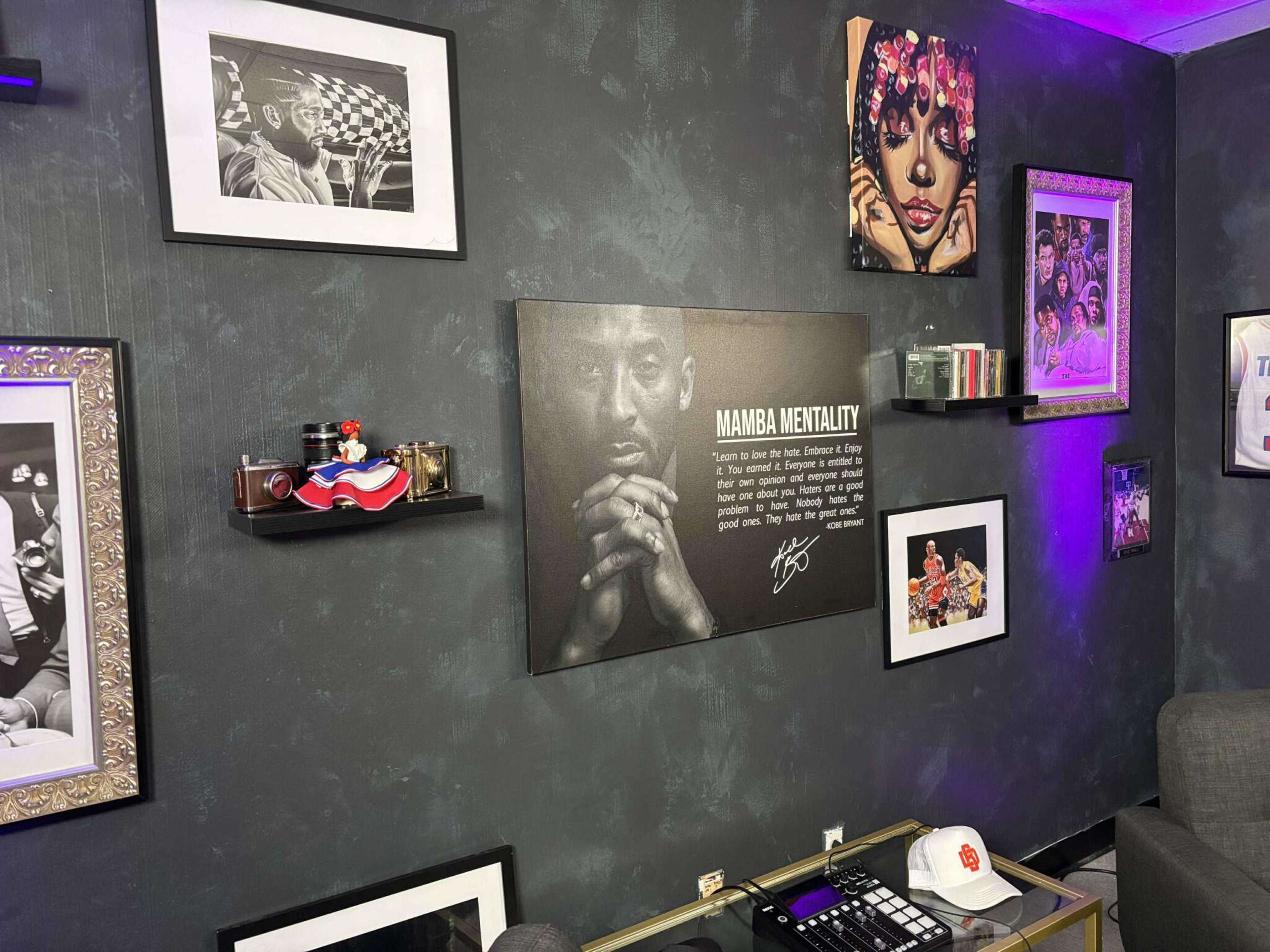 The image shows a dark wall adorned with framed photographs, shelves displaying decorative items, and a prominent artwork featuring the phrase "Mamba Mentality."