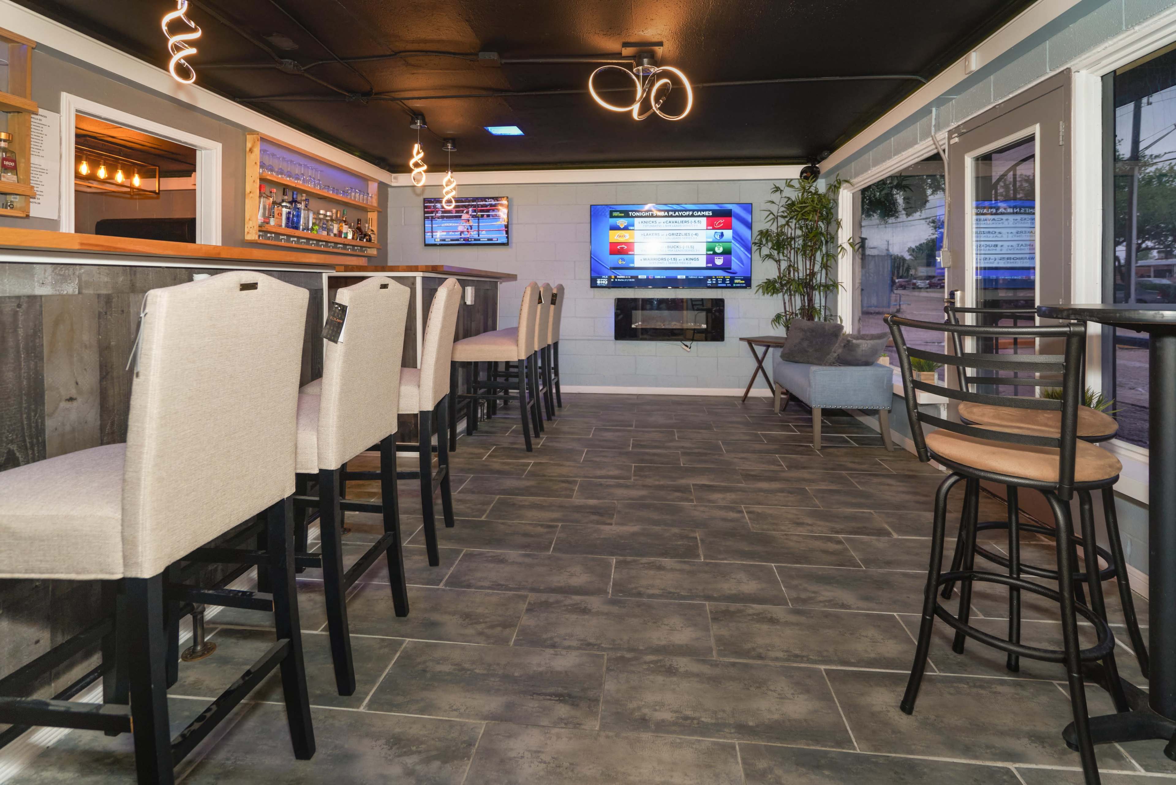 A modern bar area with high stools lining a counter, a television mounted on the wall, and a fireplace in a well-lit setting.