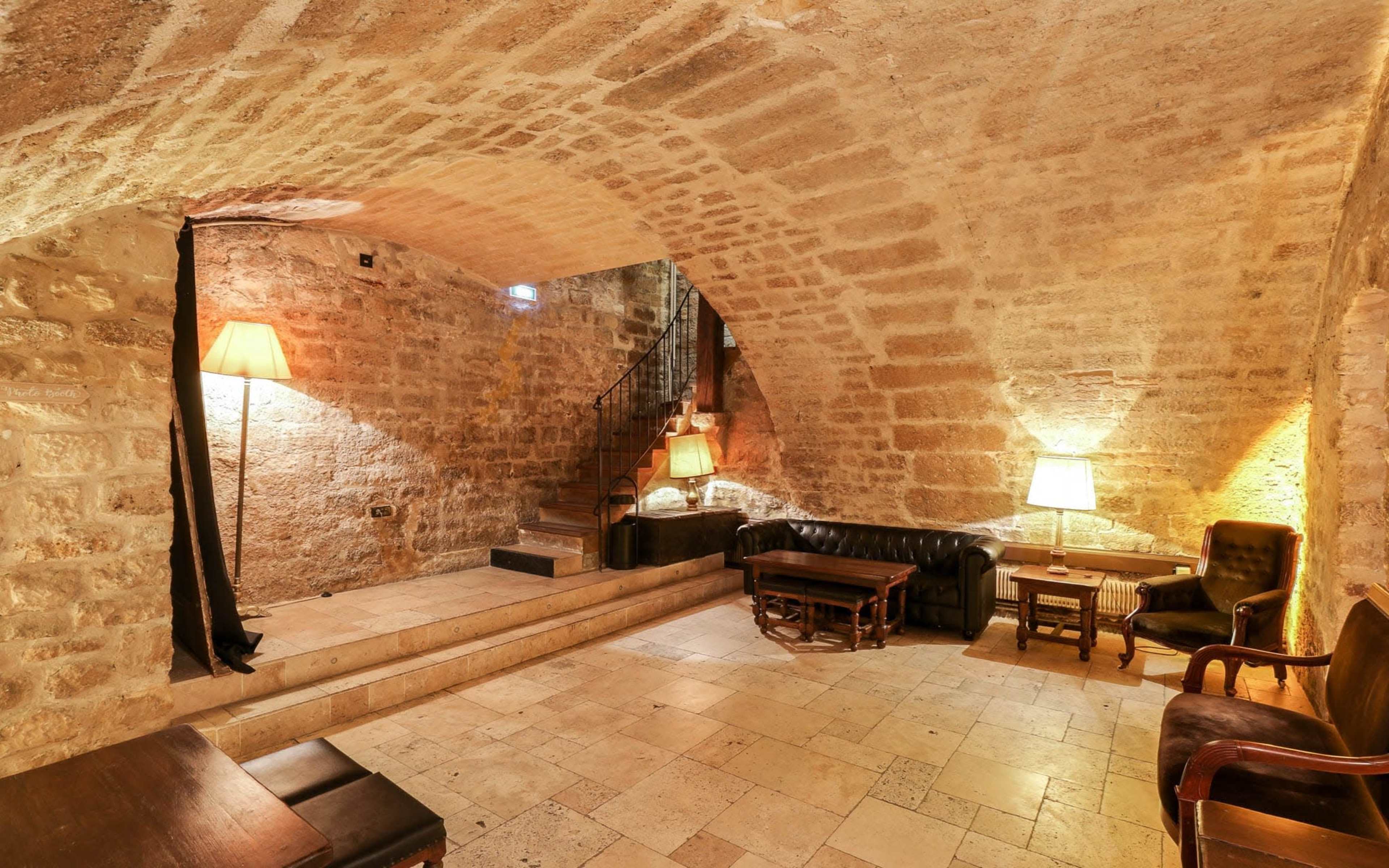 A stone-walled basement lounge with two lamps, a leather sofa, wooden tables, and a staircase leading up.