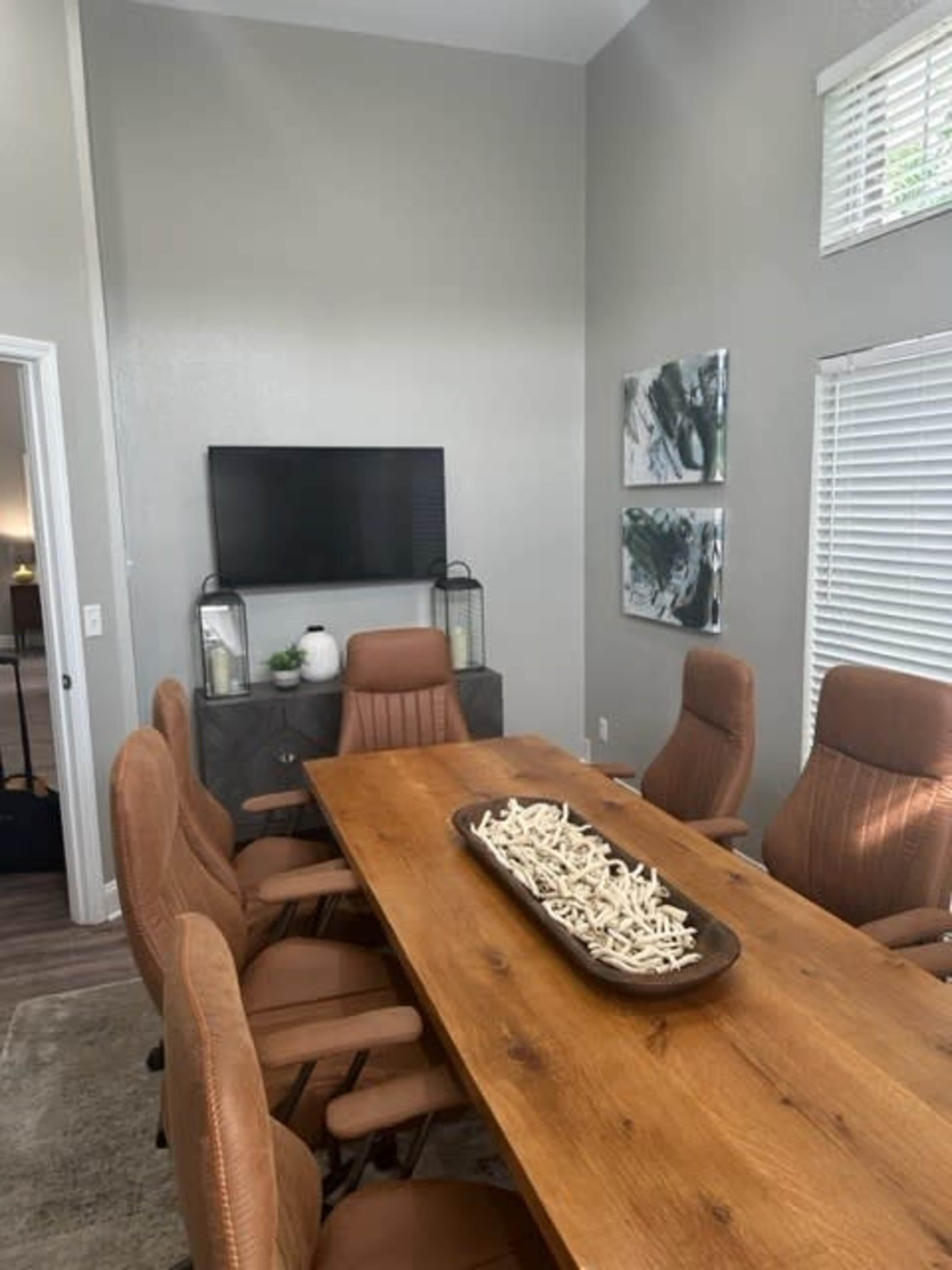 A modern conference room features a large wooden table surrounded by brown leather chairs, with a television mounted on the wall and decorative art on display.