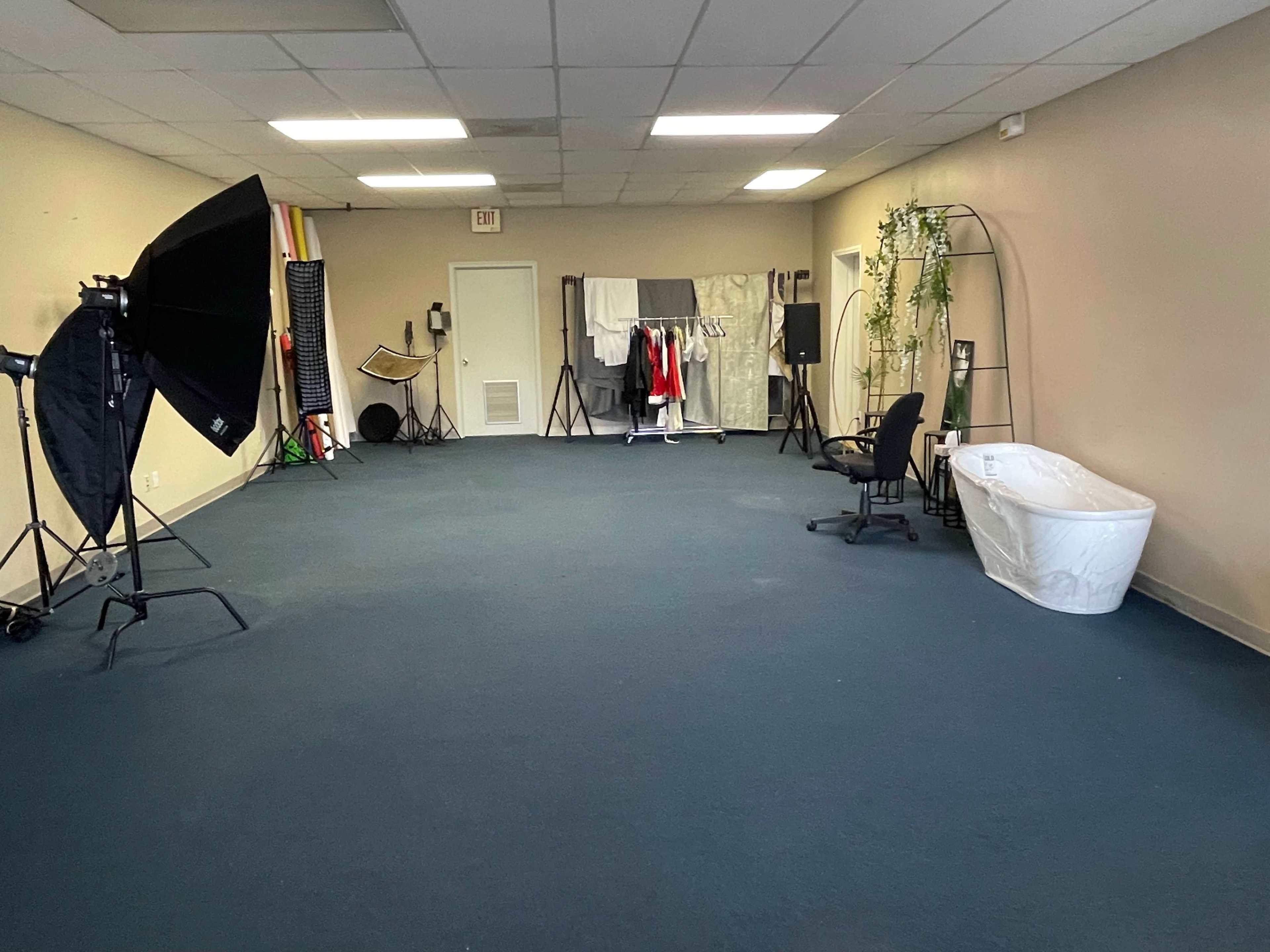 A spacious room with a blue carpet, equipped with photography lighting, a clothes rack, a chair, and a bathtub.