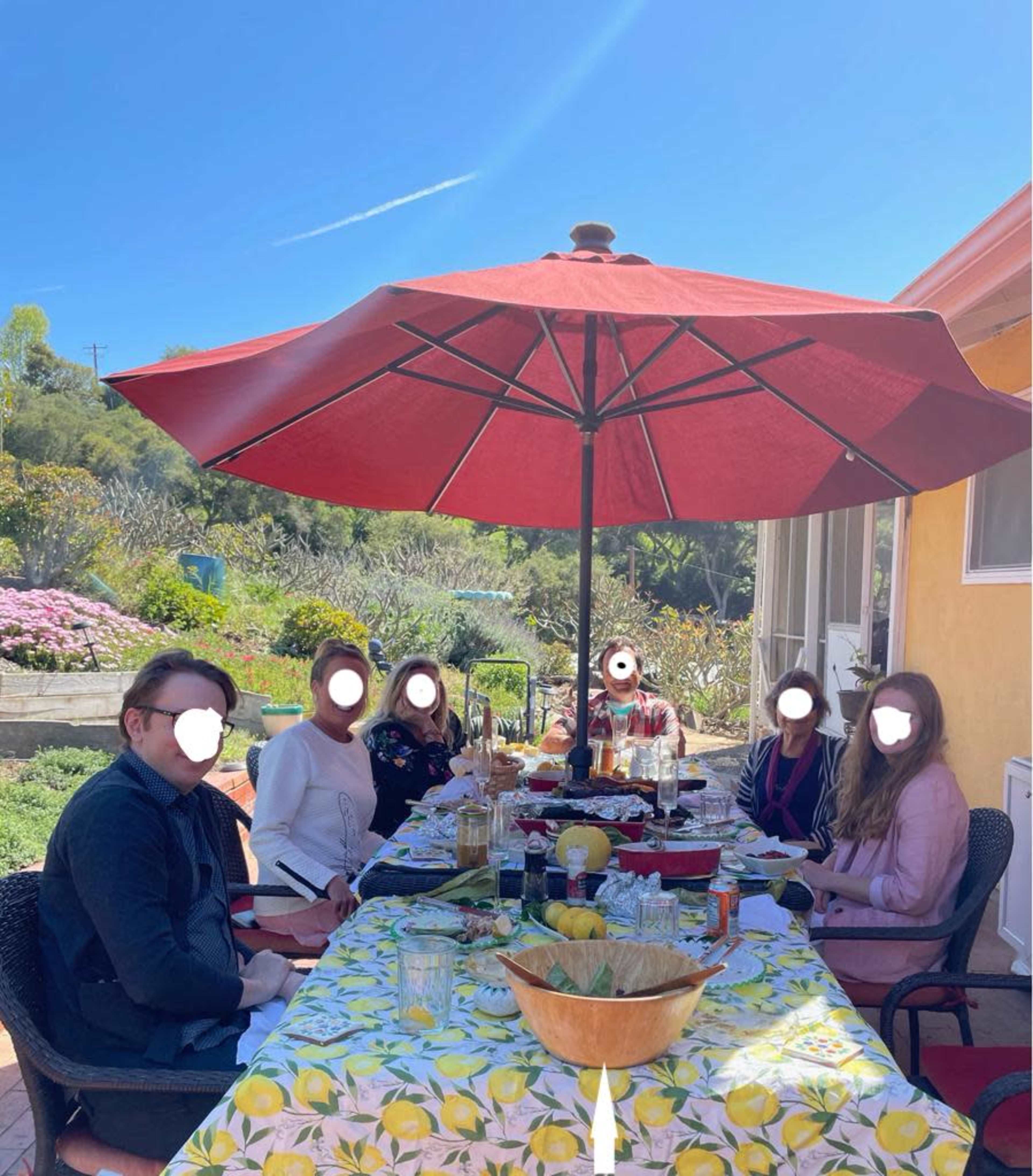 A group of six people sits around a table under a red umbrella, enjoying a meal outdoors in a sunny garden setting.
