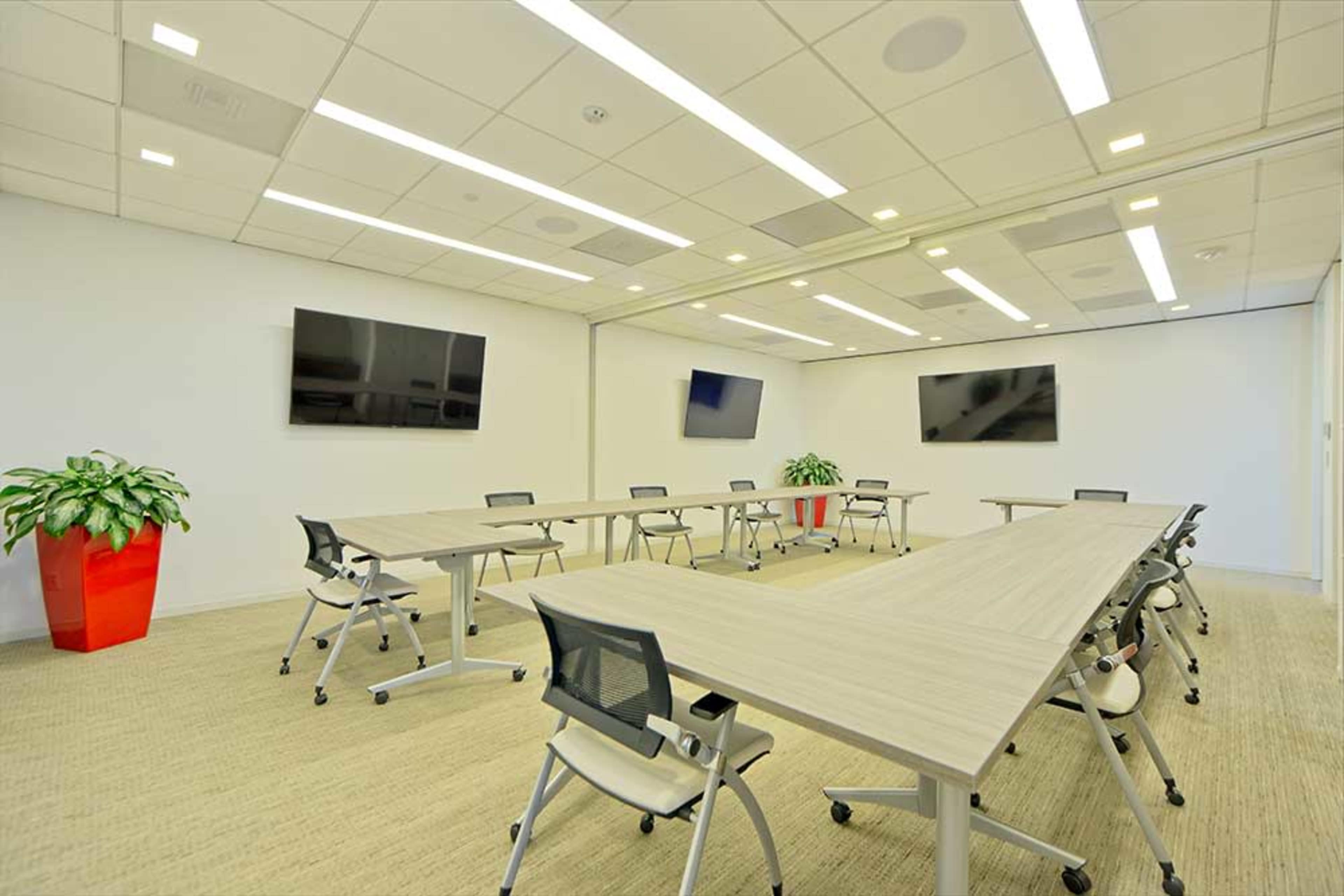 A modern conference room with multiple tables arranged in a U-shape, several televisions mounted on the walls, and a large potted plant in the corner.