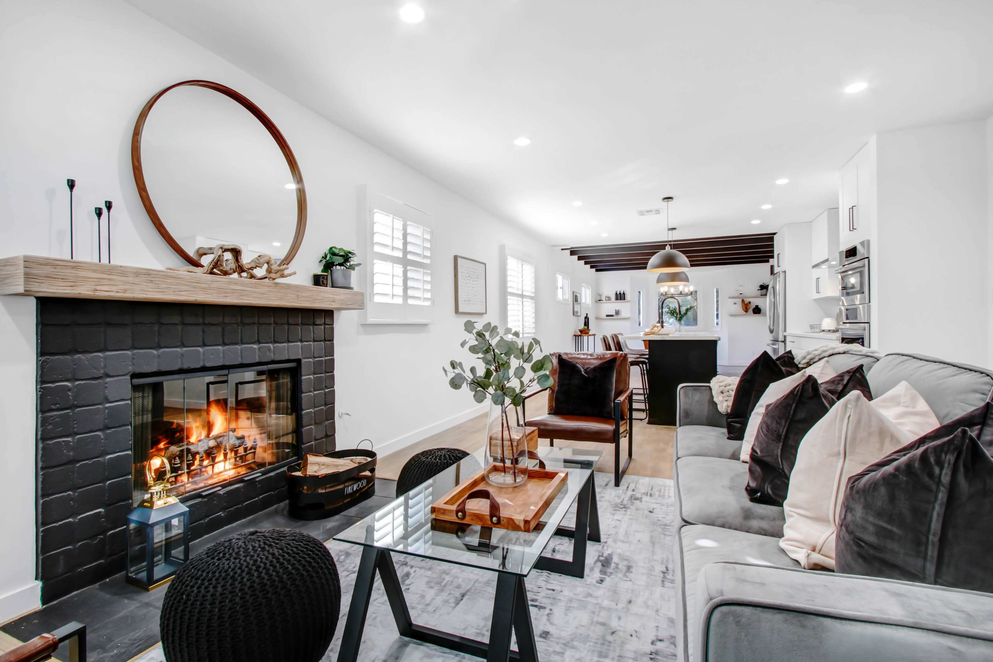 A modern living room features a black fireplace with a mirror above, a coffee table, and a cozy seating area near a kitchen with bar seating.