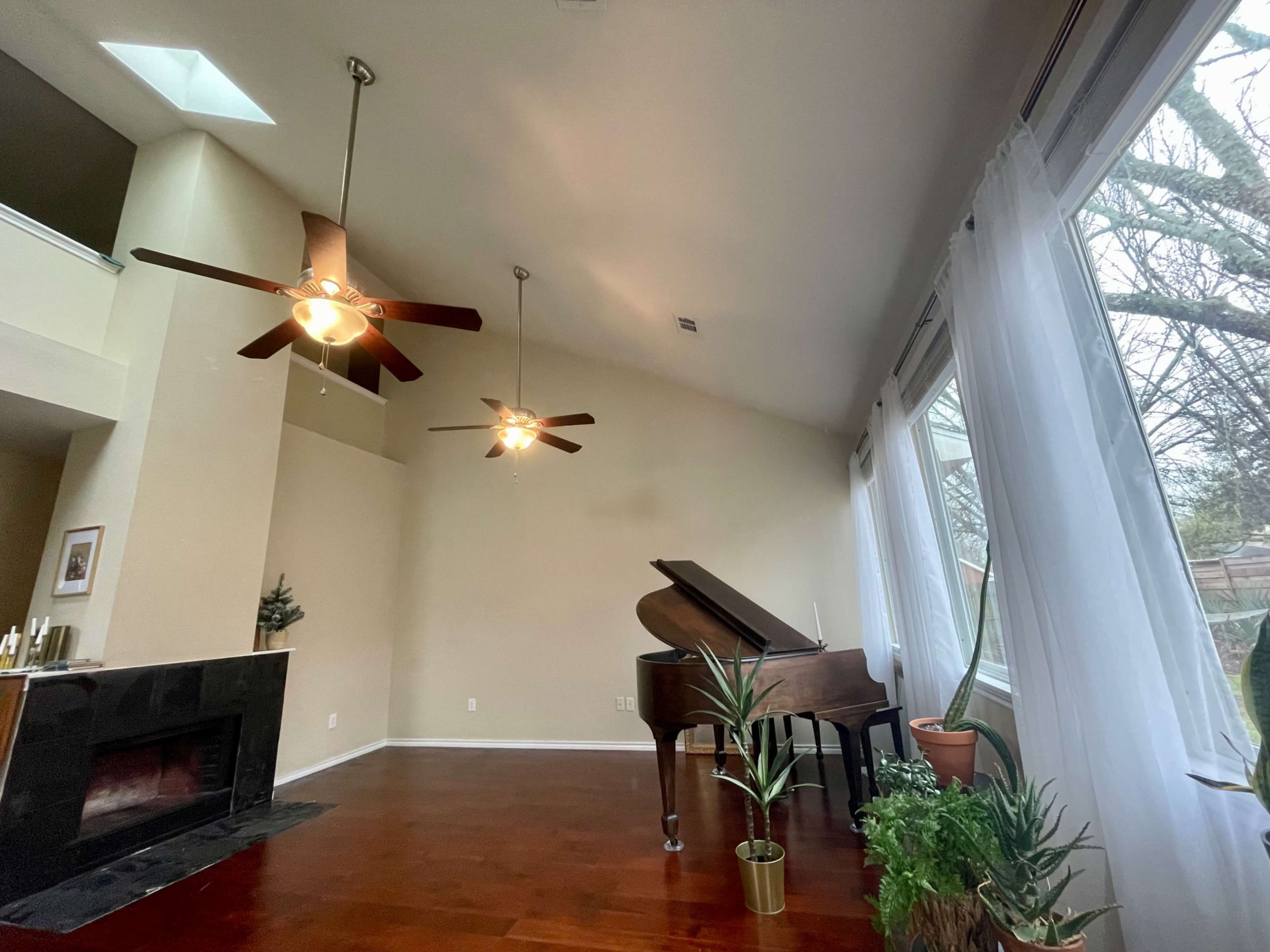 A spacious room features a grand piano, two ceiling fans, and large windows with sheer curtains overlooking a garden.