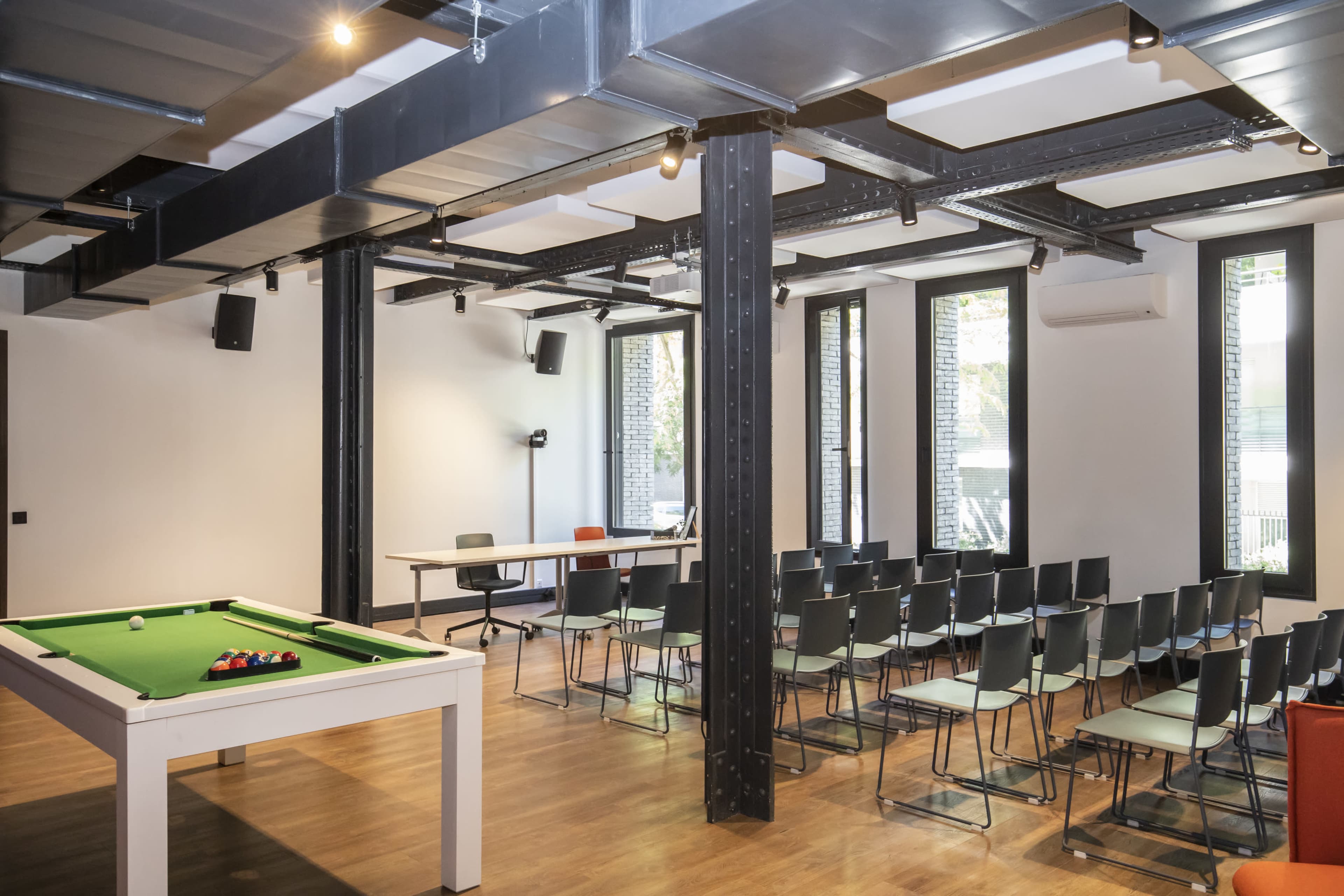 A spacious room features a pool table on one side and a row of chairs facing a table on the other, with large windows providing natural light.