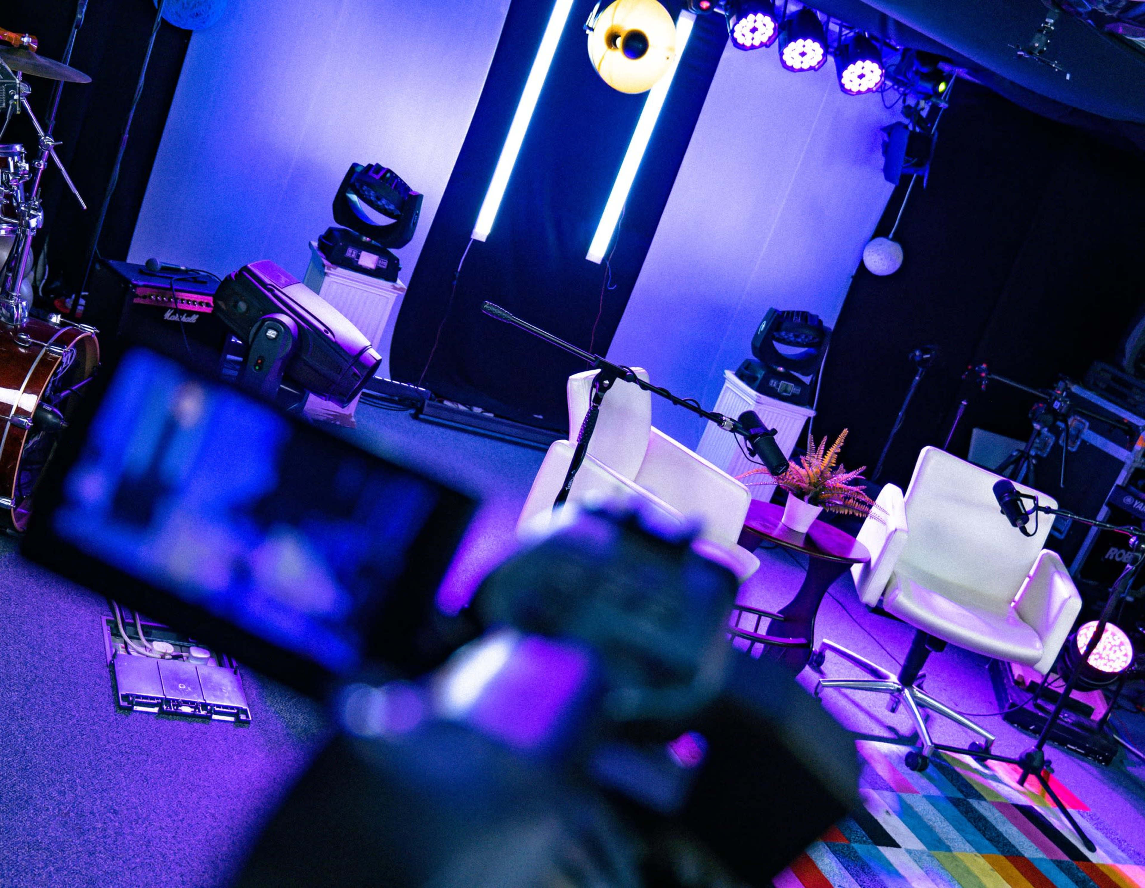 The image shows a brightly lit studio setup featuring two white chairs placed in front of sound equipment and colorful striped flooring.
