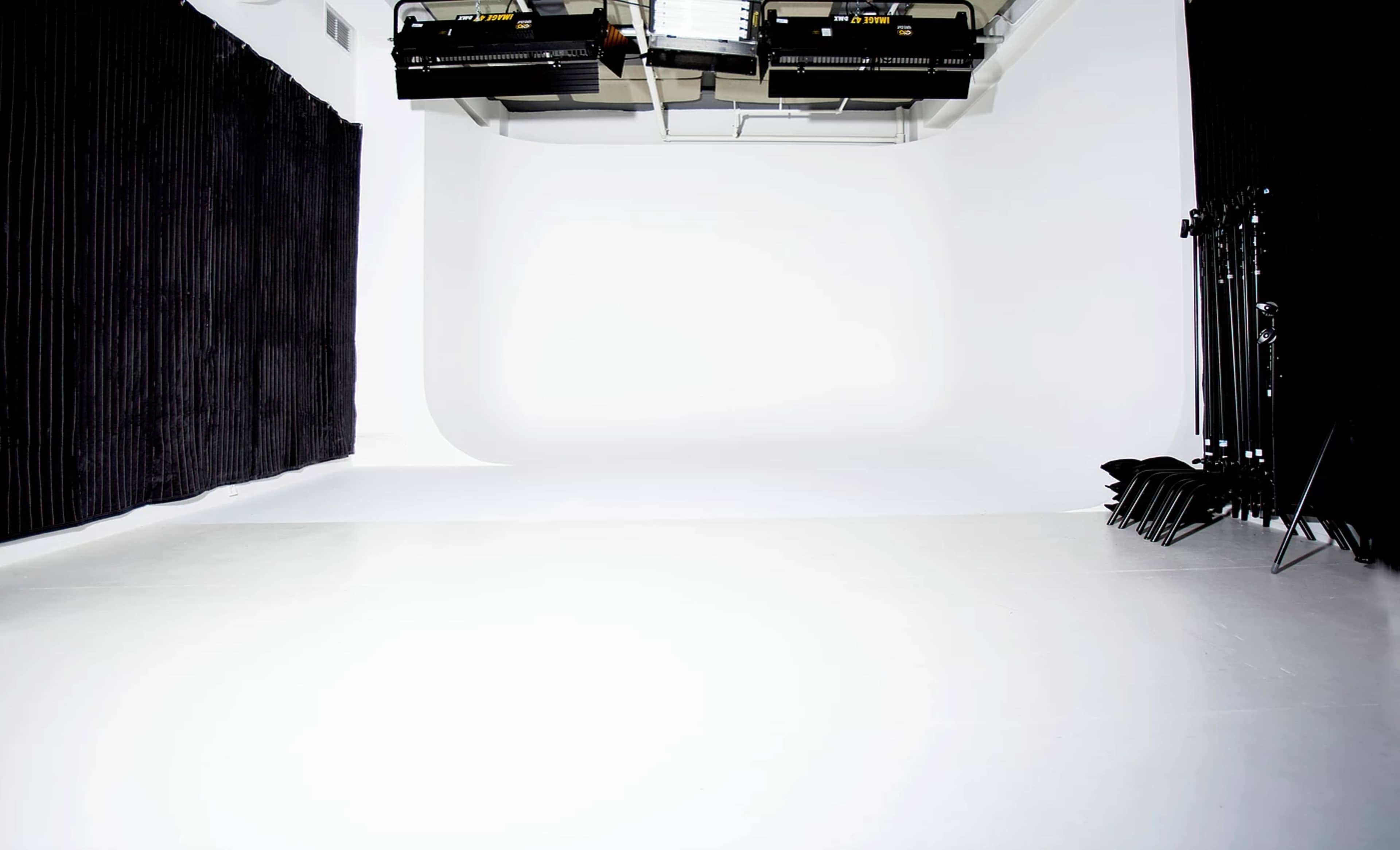 The image shows a photo studio with a seamless white backdrop, black curtains, and lighting equipment mounted on the ceiling.