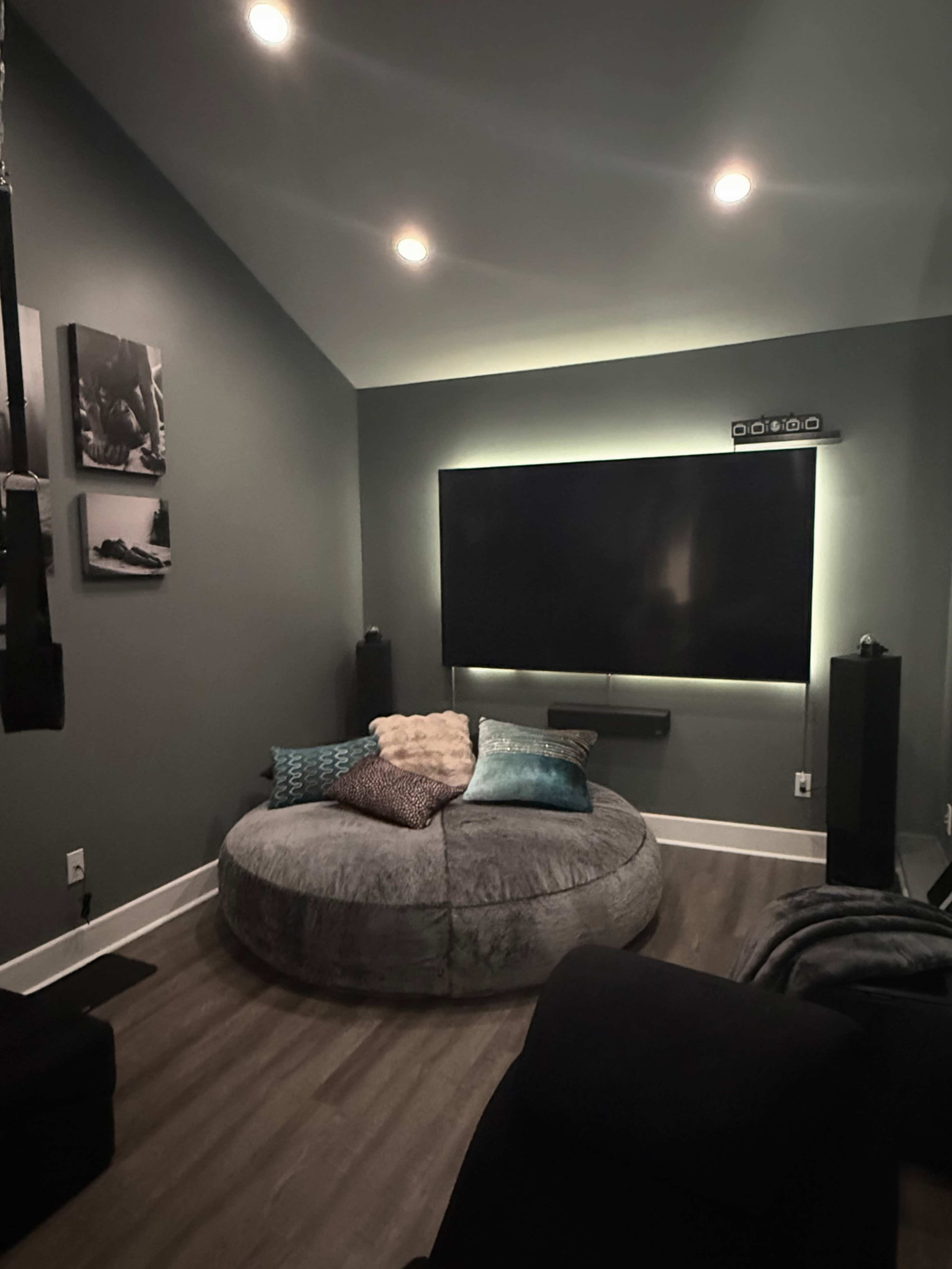 The image shows a cozy home theater setup featuring a round, plush seating area in front of a large wall-mounted television, with soft lighting and wall decor.