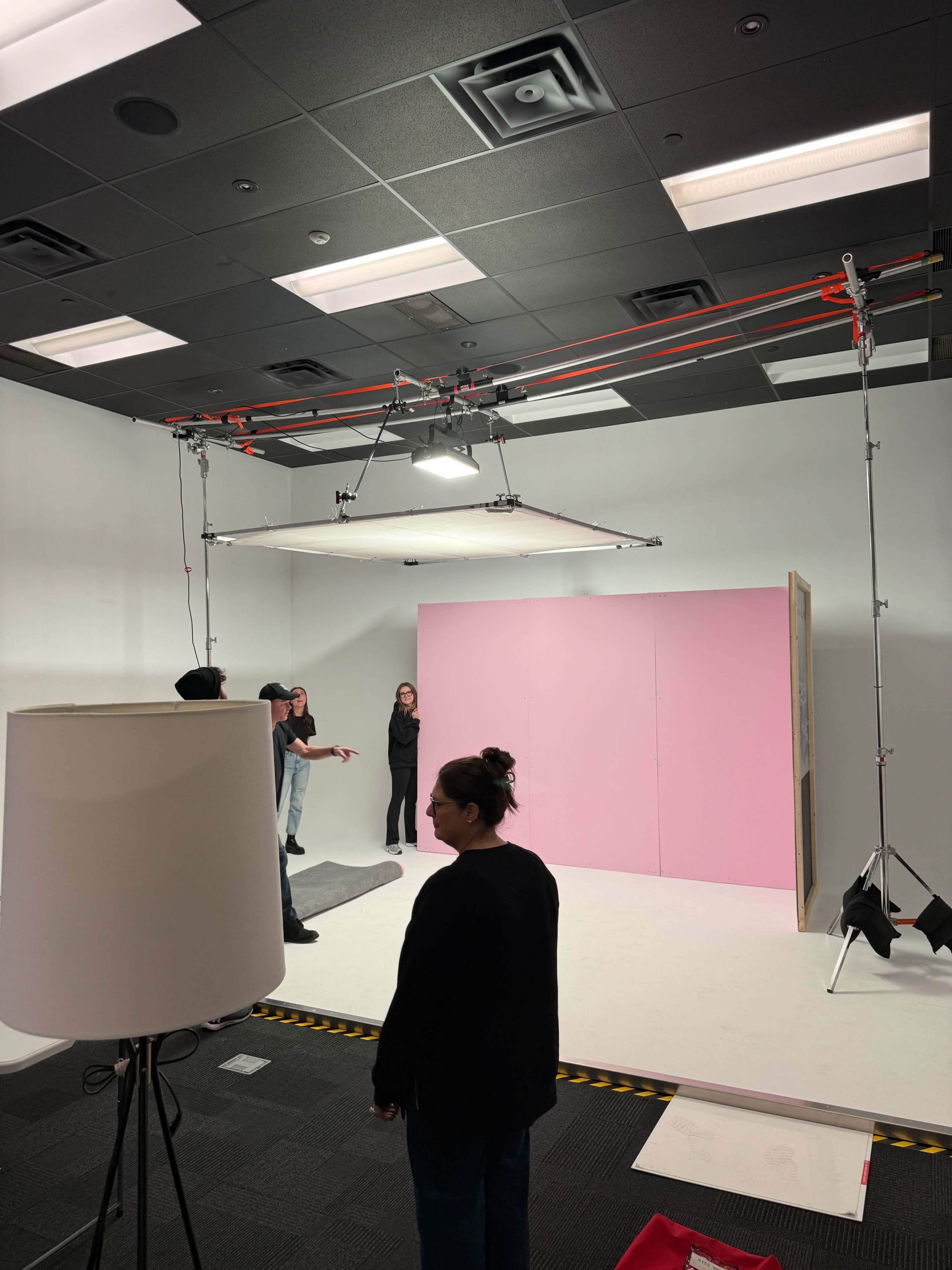 A film crew sets up a shoot in a studio, featuring a pink backdrop and various lighting equipment.