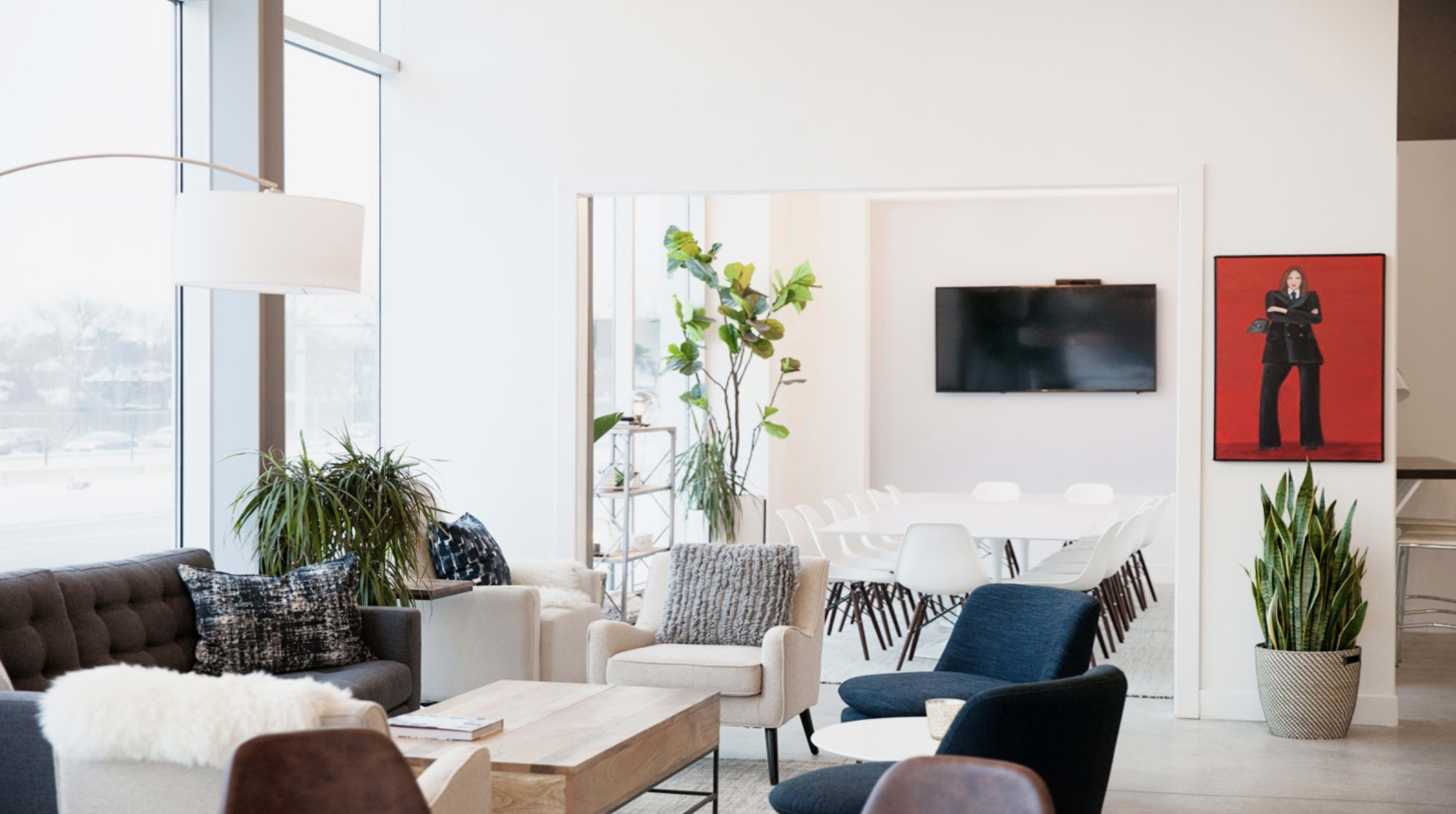 The image shows a modern, minimalist interior with a mix of seating areas, plants, and a view of a meeting room featuring a television and artwork on the wall.