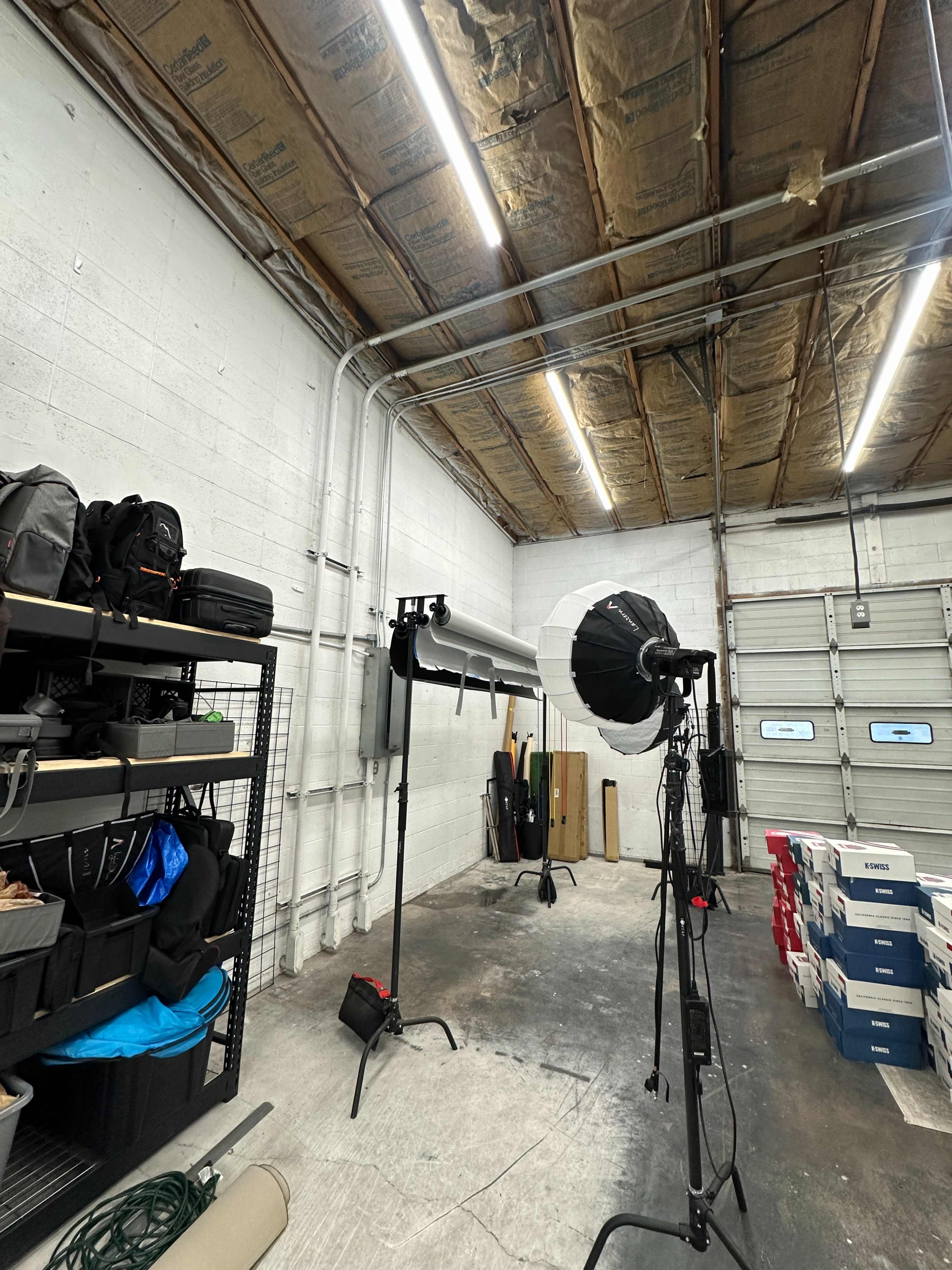 A well-lit photography studio features a seamless backdrop, lighting equipment, and shelves stacked with various bags and supplies.
