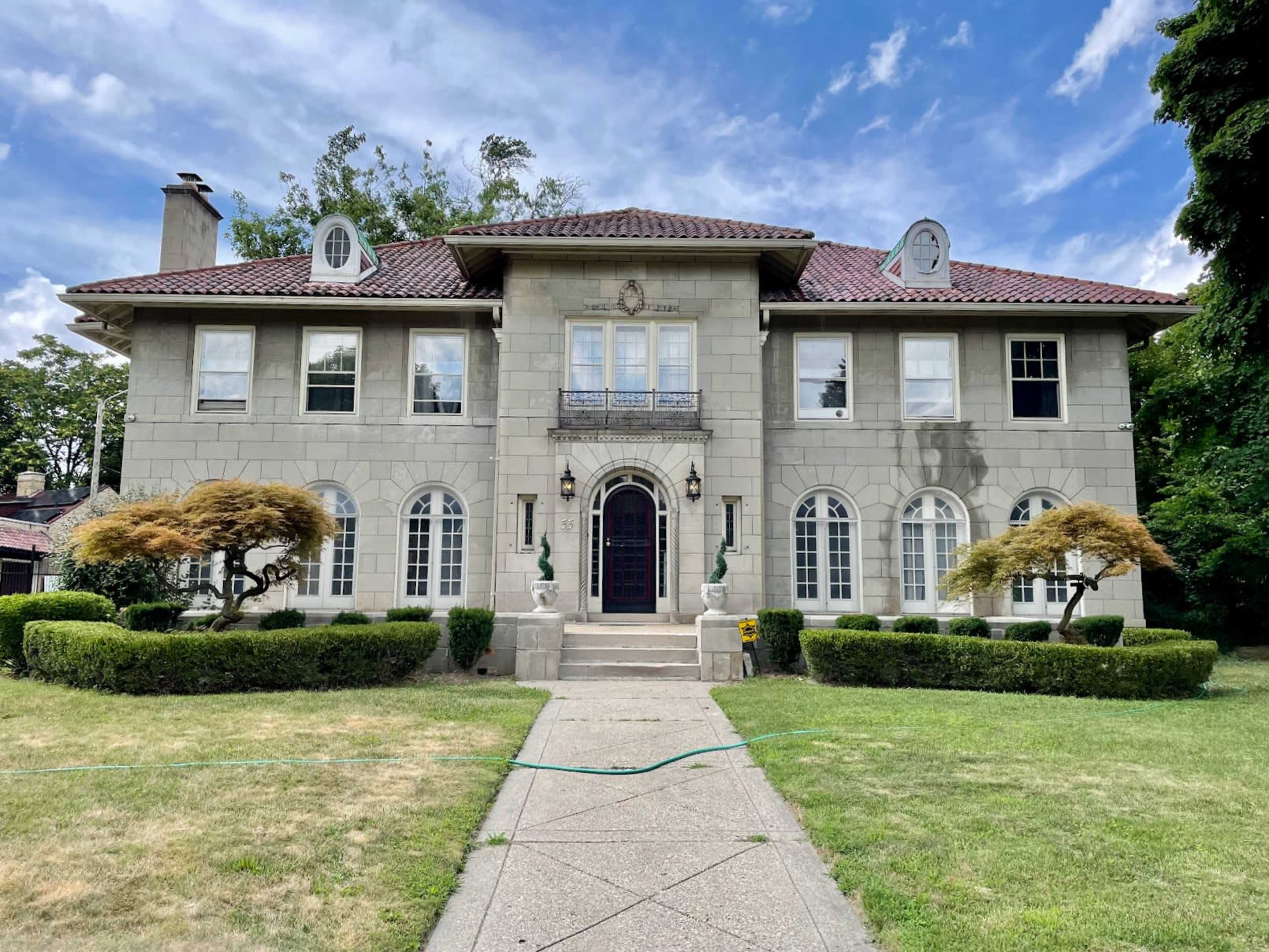 A large, two-story stone house with a tiled roof, flanked by manicured shrubs and two ornamental trees in the front yard.