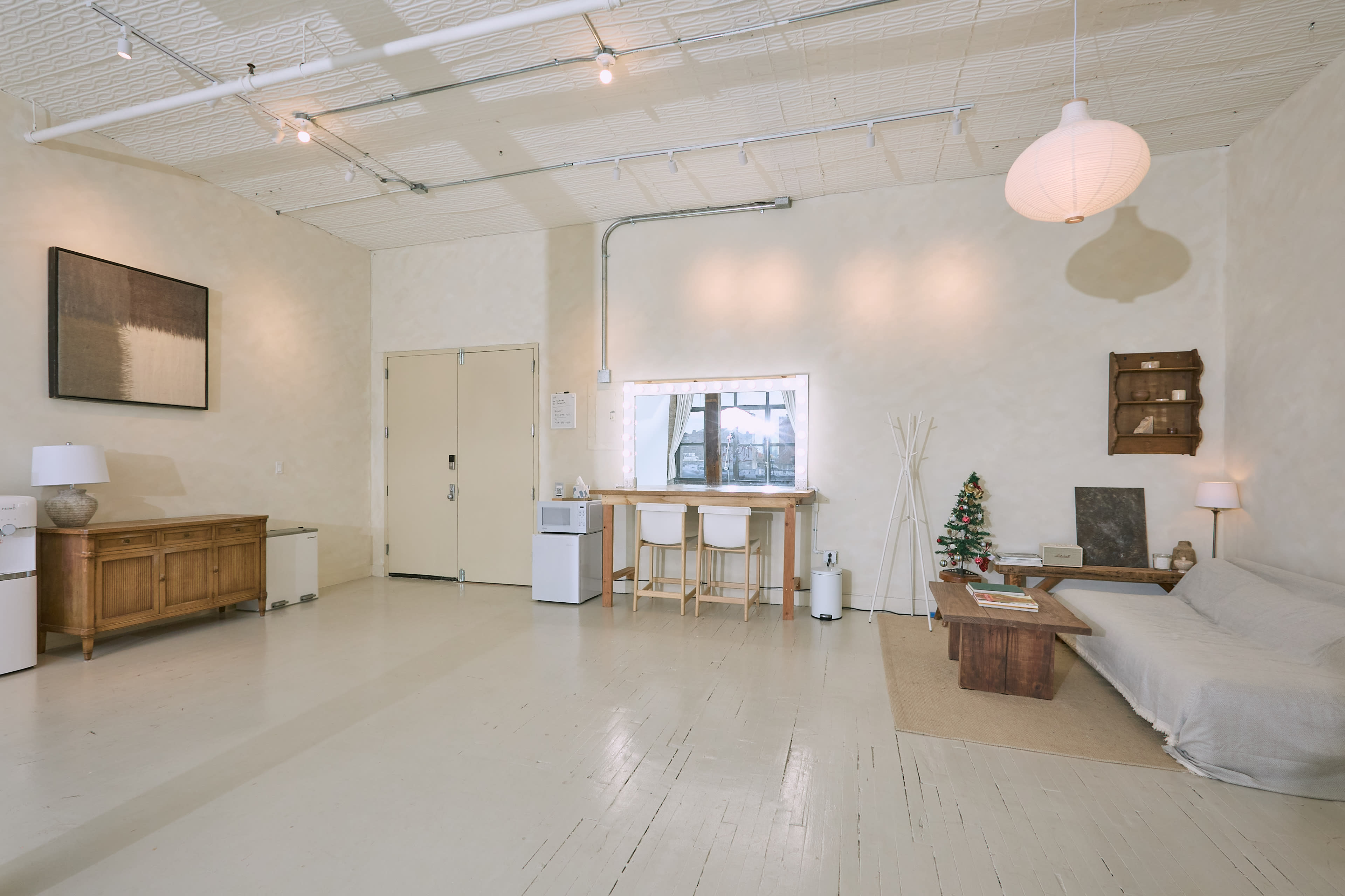 The image shows a minimalist, light-colored interior space featuring a couch, a small dining area with stools, and various decorative elements along the walls.
