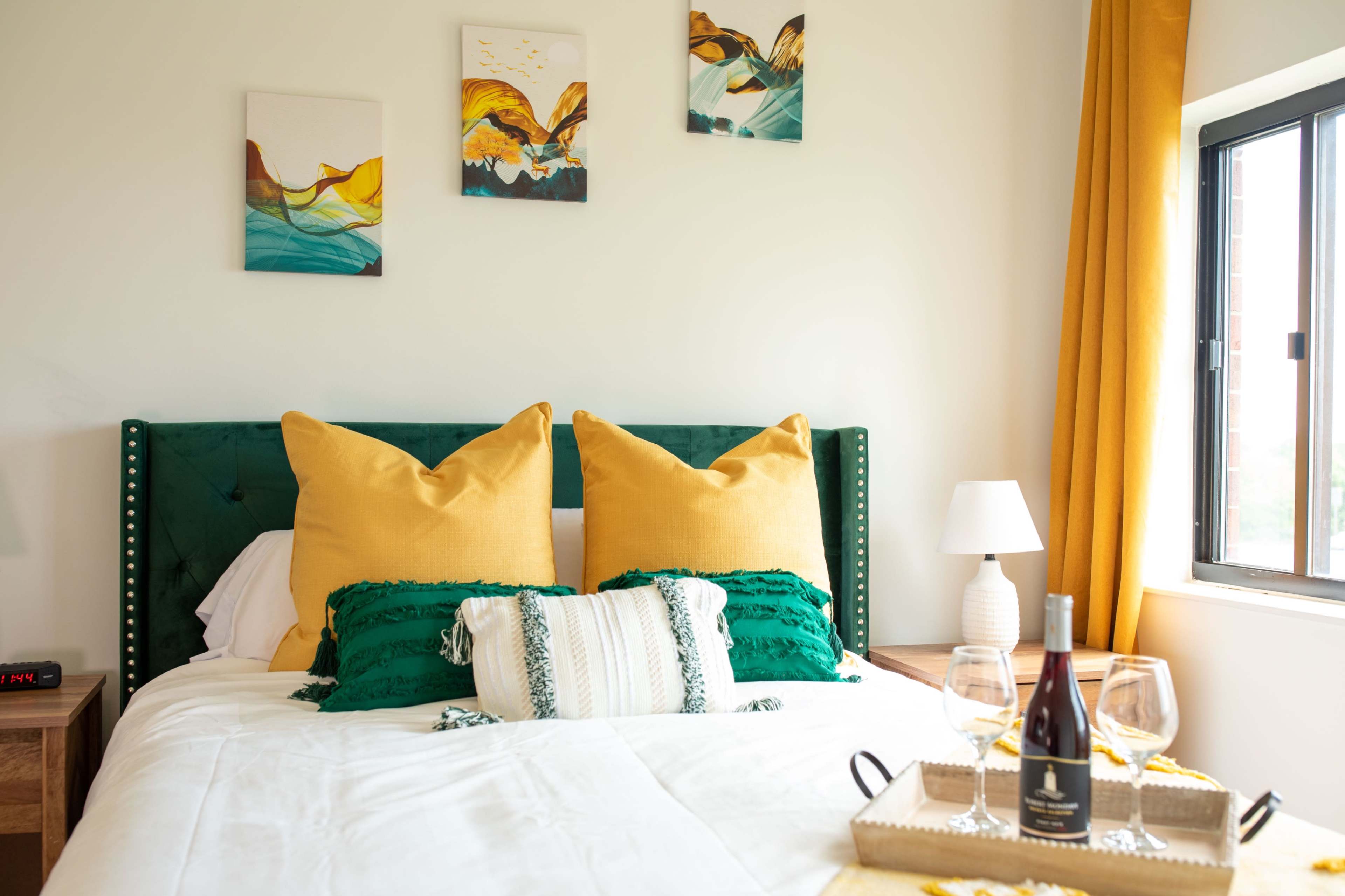 A neatly made bed features vibrant yellow pillows and a decorative throw, with a tray holding a bottle of wine and glasses set beside a window with yellow curtains.