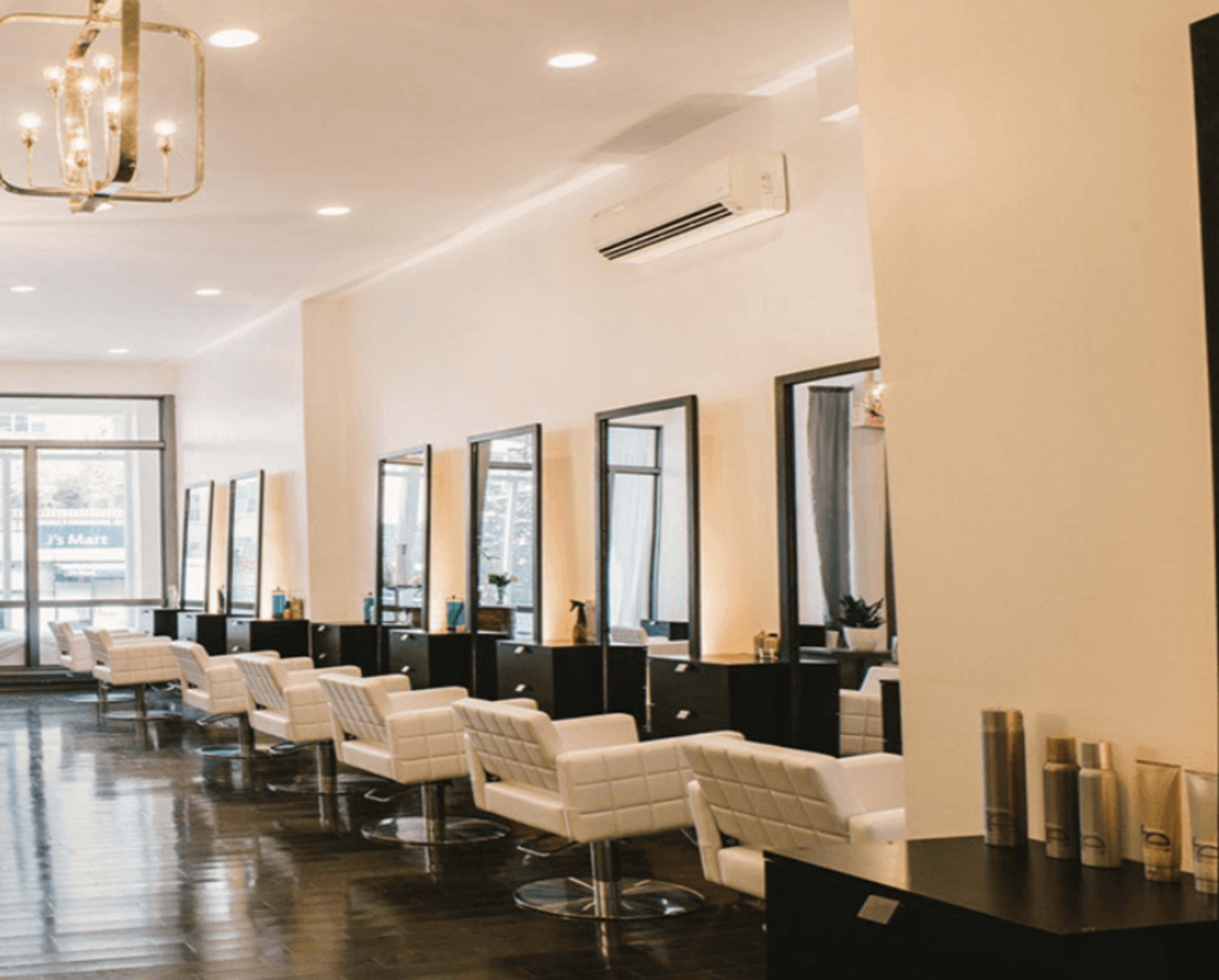 The image shows a modern hair salon with several styling stations, each equipped with mirrors and white chairs, lined along a well-lit interior.