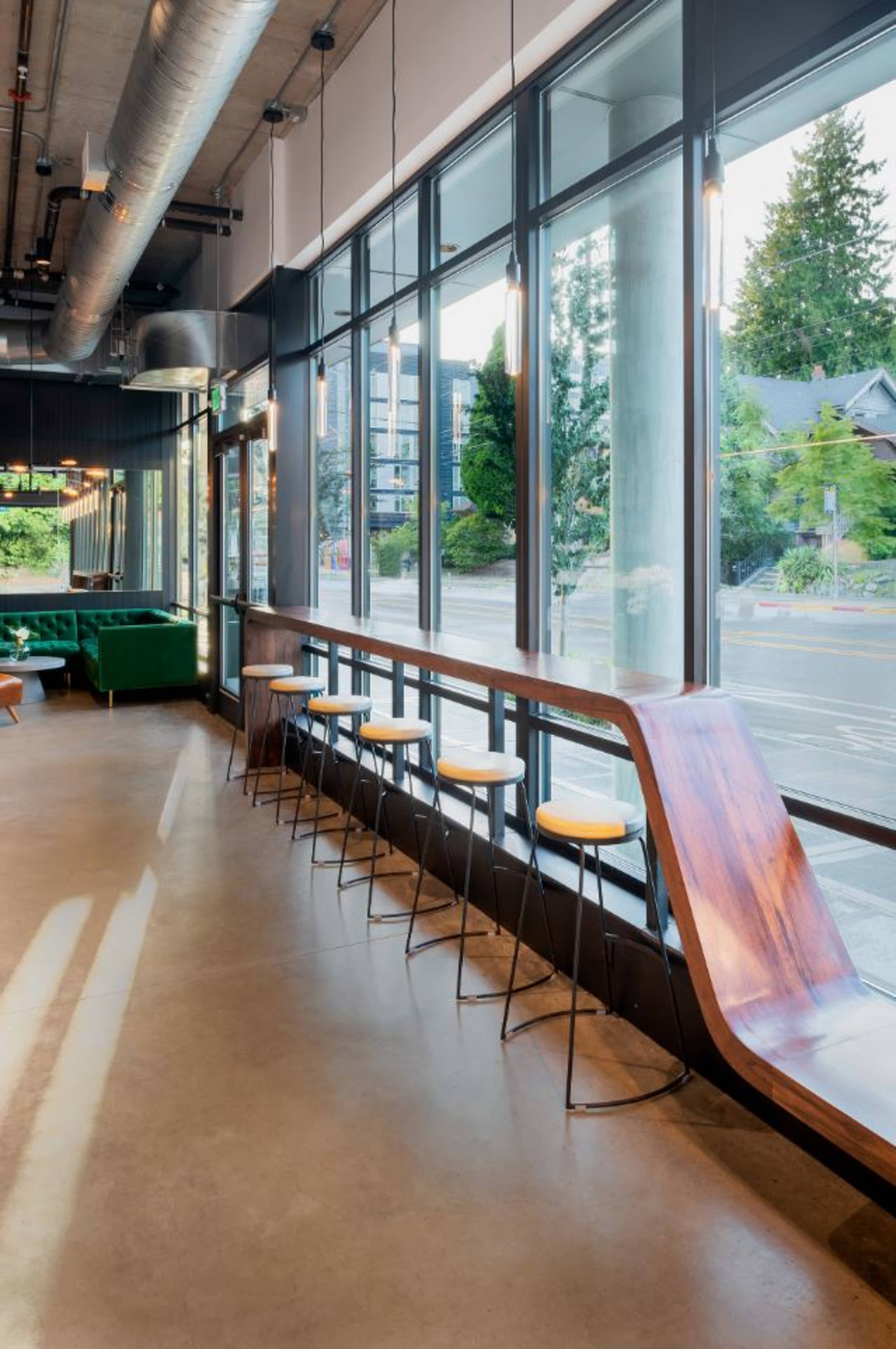 A modern café interior featuring a woodenCounter with three stools, large windows allowing natural light, and a view of trees outside.