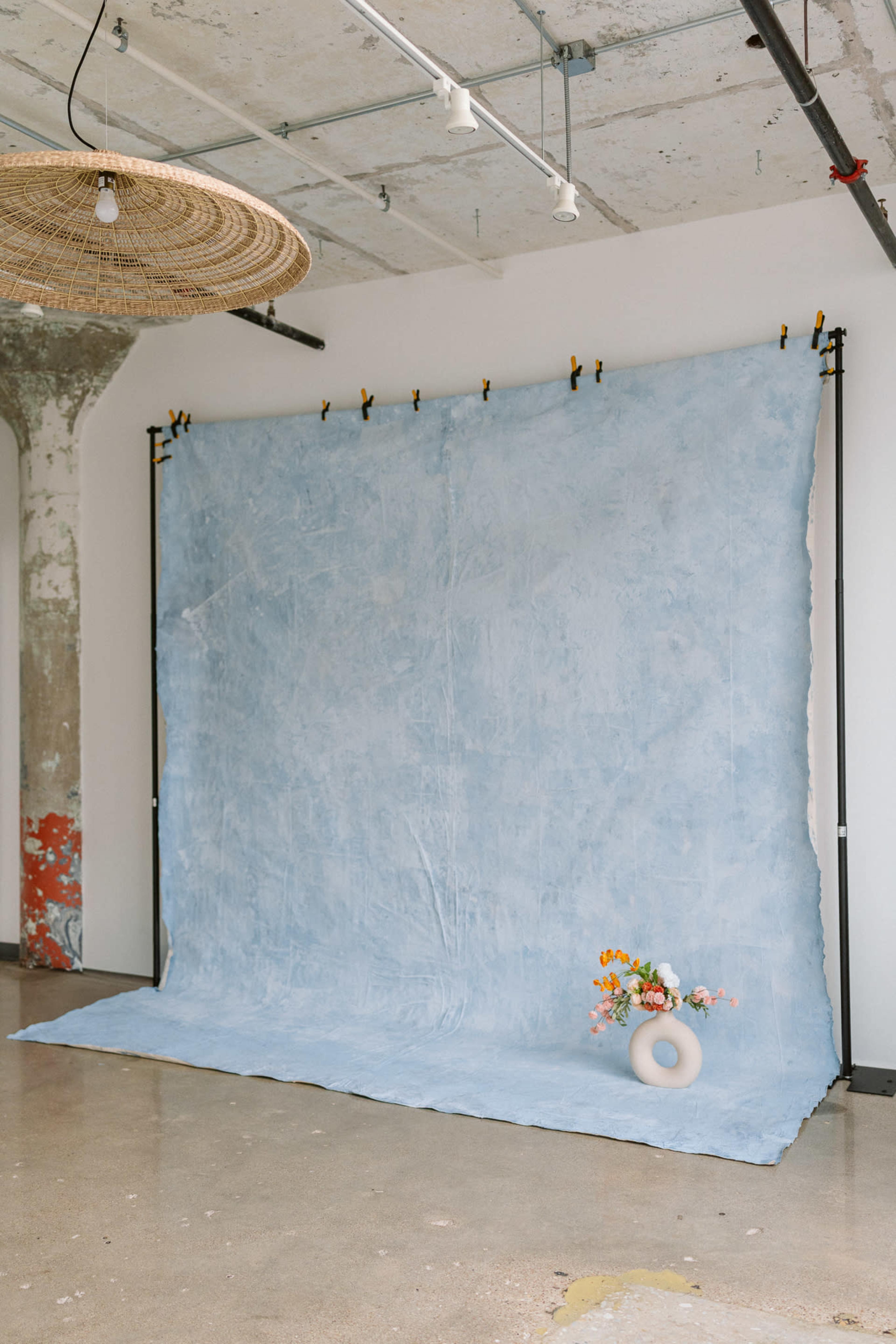 A light blue backdrop is set up in a spacious room with a polished concrete floor, featuring a decorative hanging light and a floral arrangement placed in front.