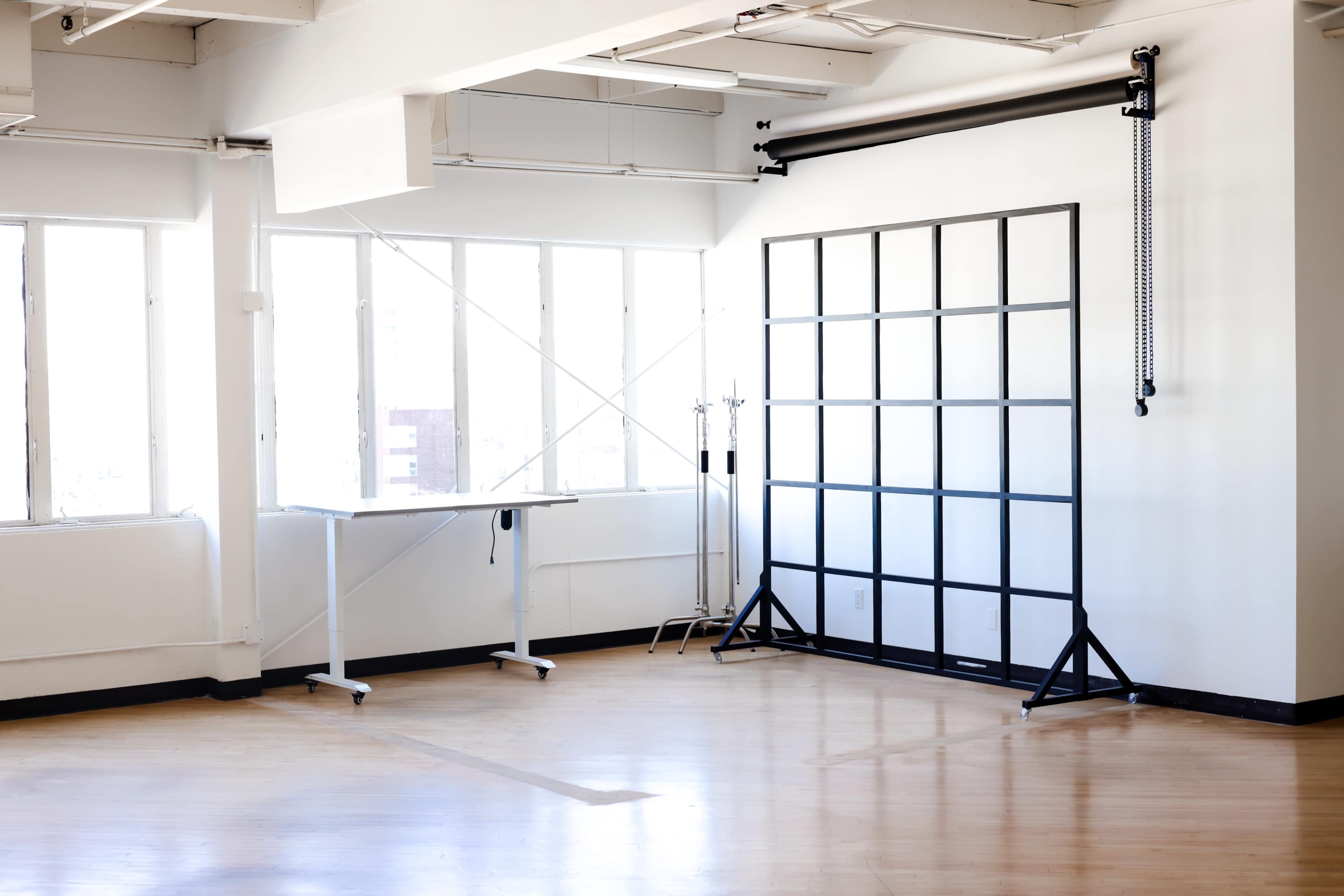 A spacious studio features large windows, a simple black backdrop setup, and a standing white table on a wooden floor.