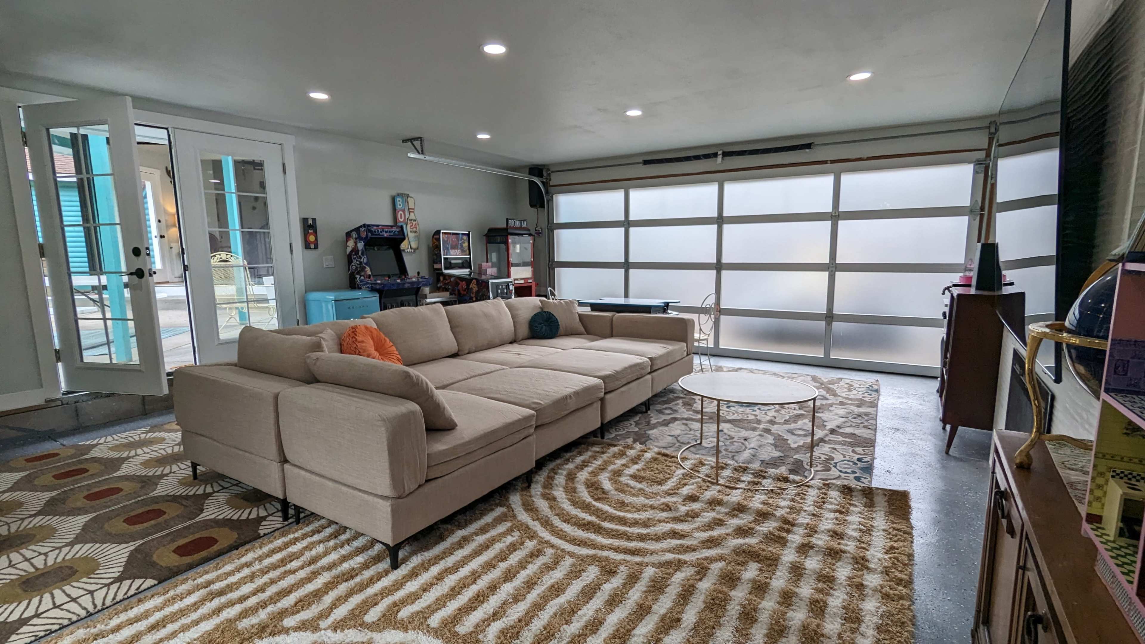 The image shows a spacious living area featuring a large beige sectional sofa, a round coffee table, and a decorative rug, with a glass garage door allowing light into the room.