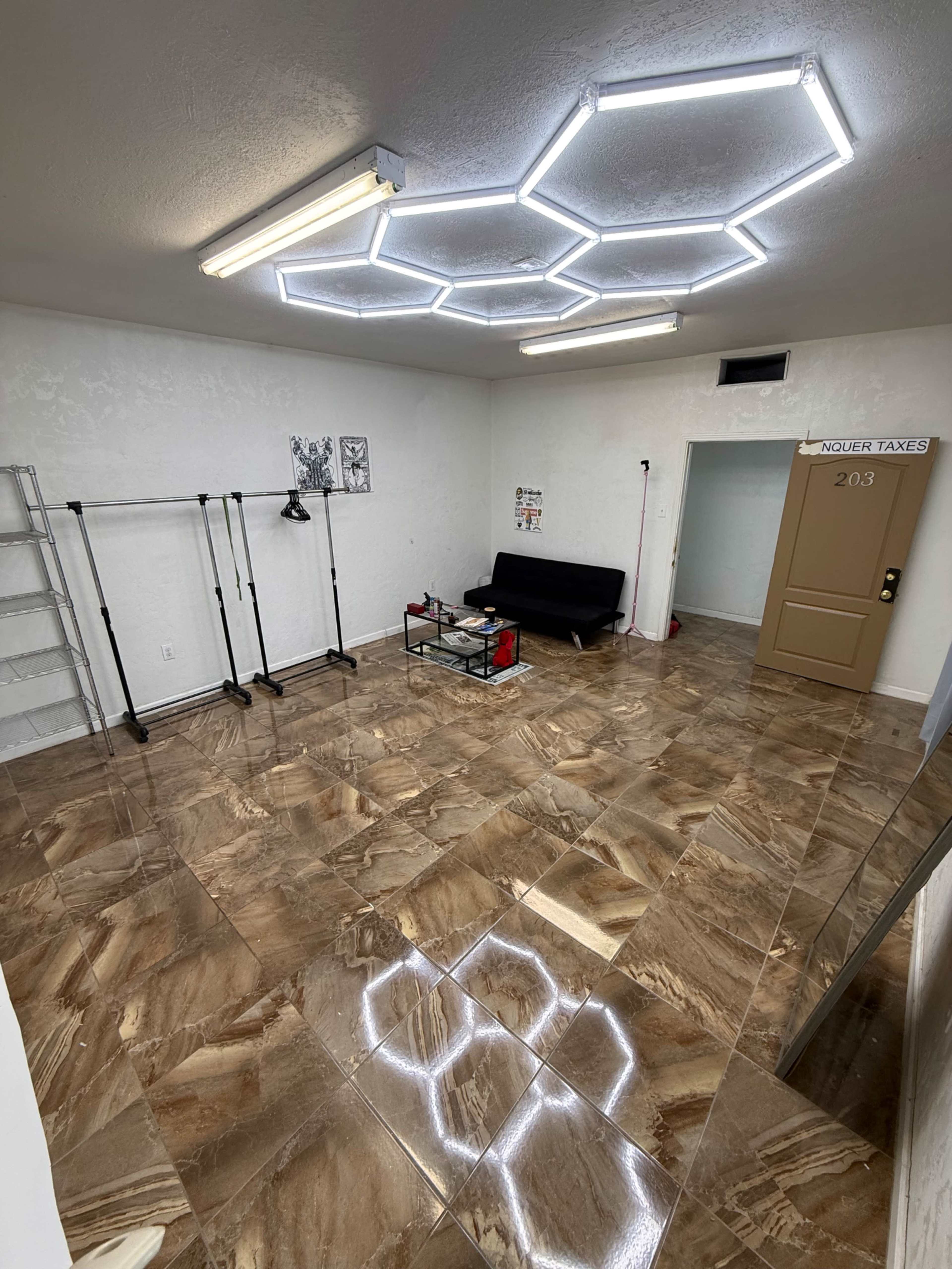 The image shows a spacious room with shiny, patterned tiles, a black couch, a glass coffee table, and several wall-mounted lights in a hexagonal arrangement on the ceiling.