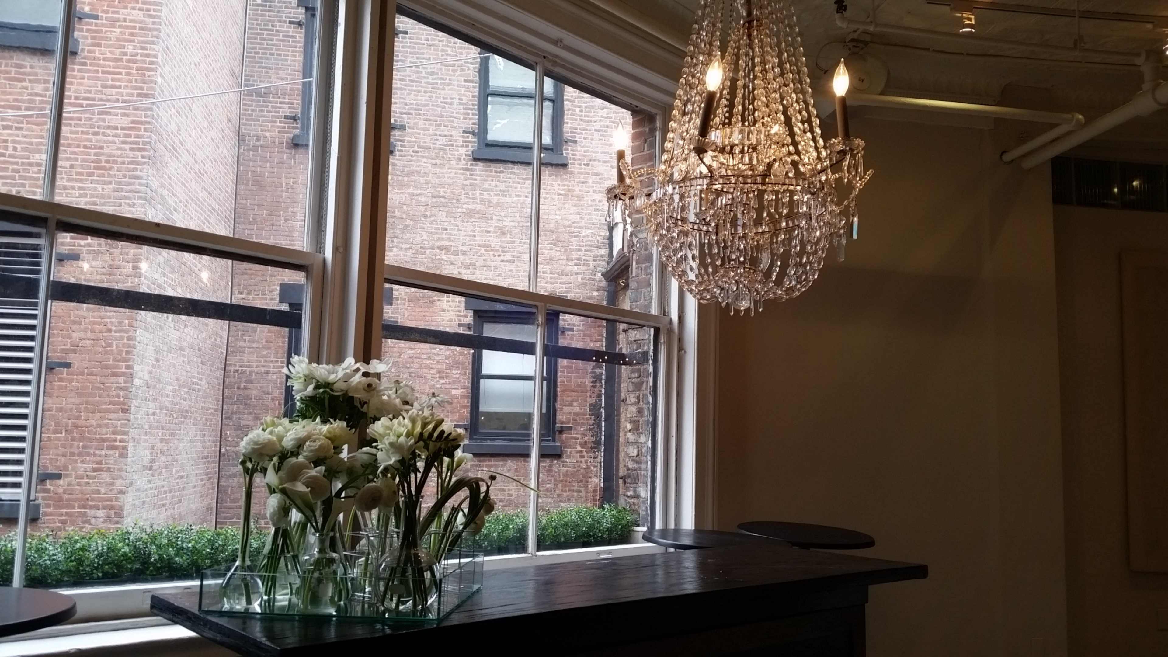 A chandelier hangs from the ceiling above a table with a vase of white flowers, set against a backdrop of brick buildings visible through the windows.