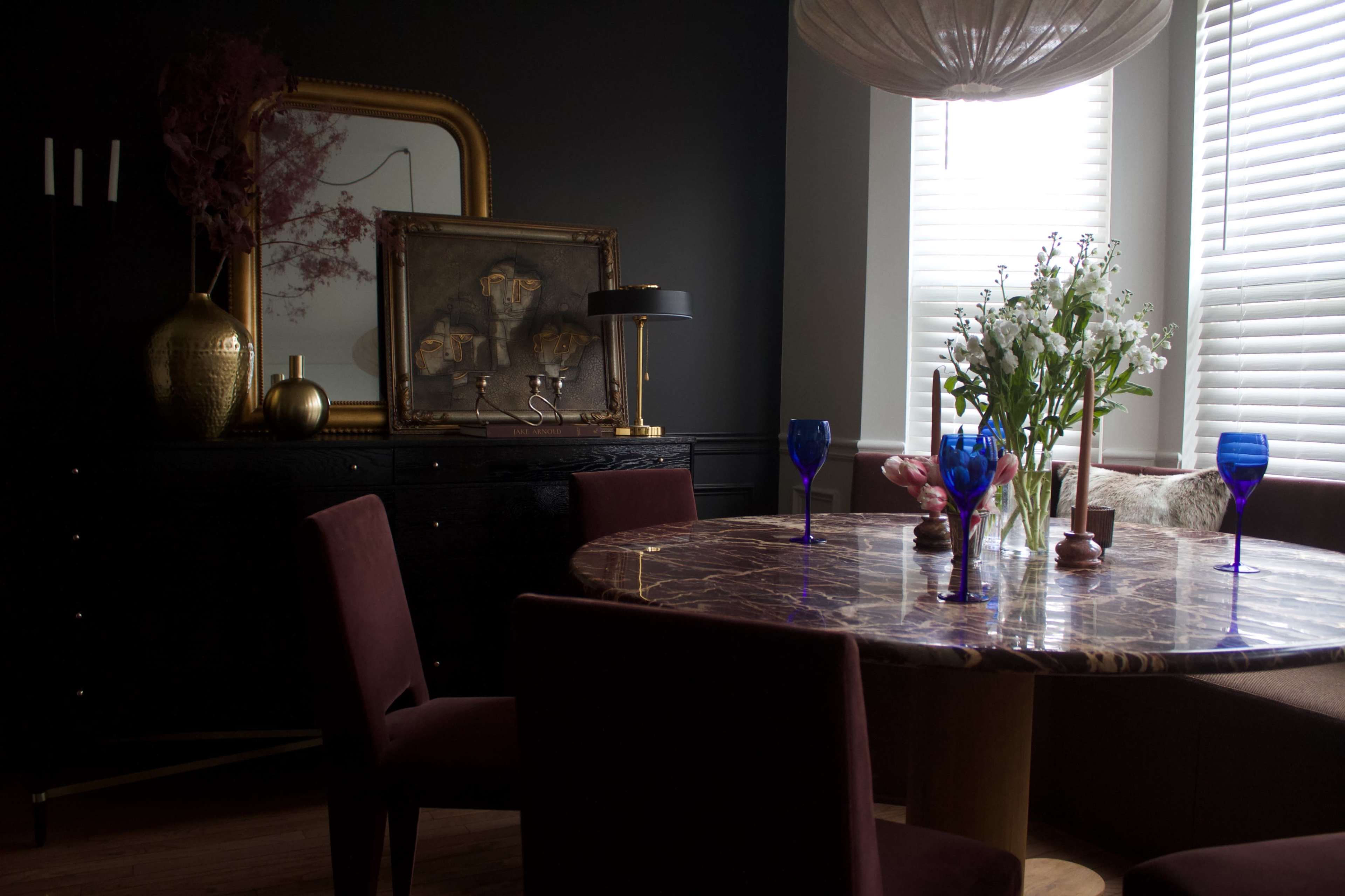 A dark room features a round marble dining table surrounded by burgundy chairs, with a vase of flowers at the center and a gold-framed artwork on the wall.