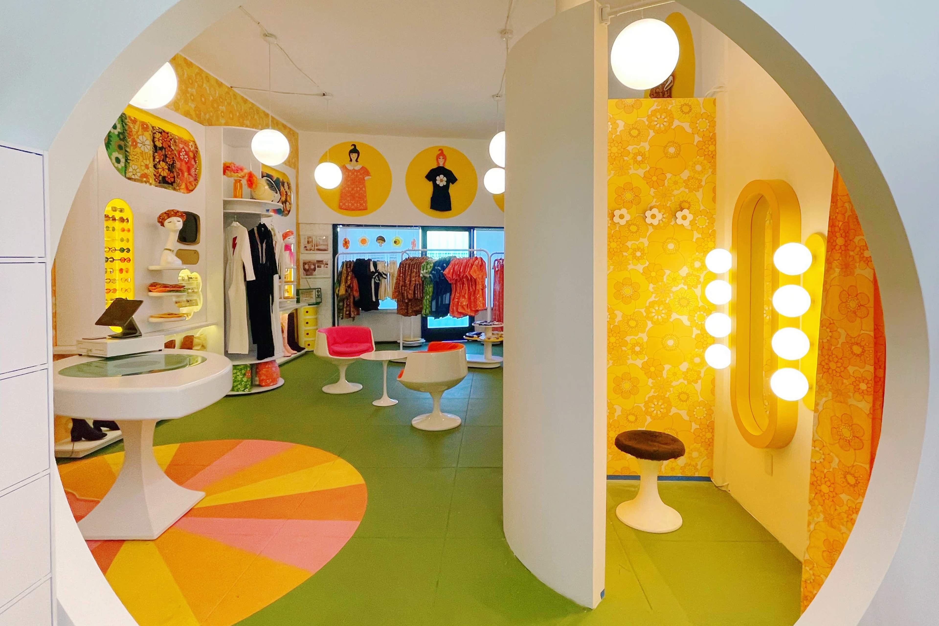 The image shows a vibrant clothing store interior with circular displays, bright yellow floral wallpaper, and retro-style furniture.