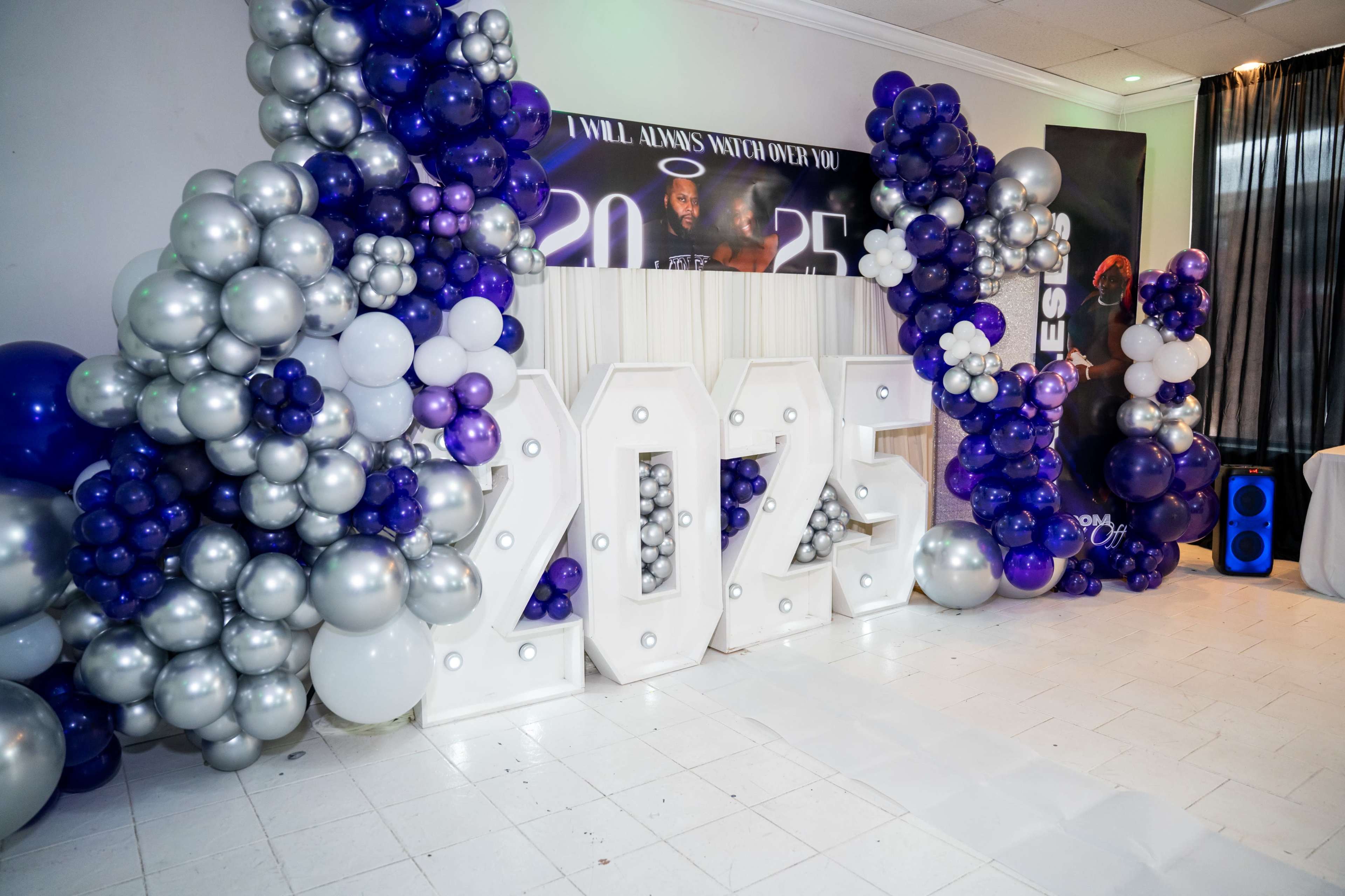 The image depicts a decorated event space featuring large, illuminated numbers "2025" surrounded by clusters of purple, silver, and white balloons.