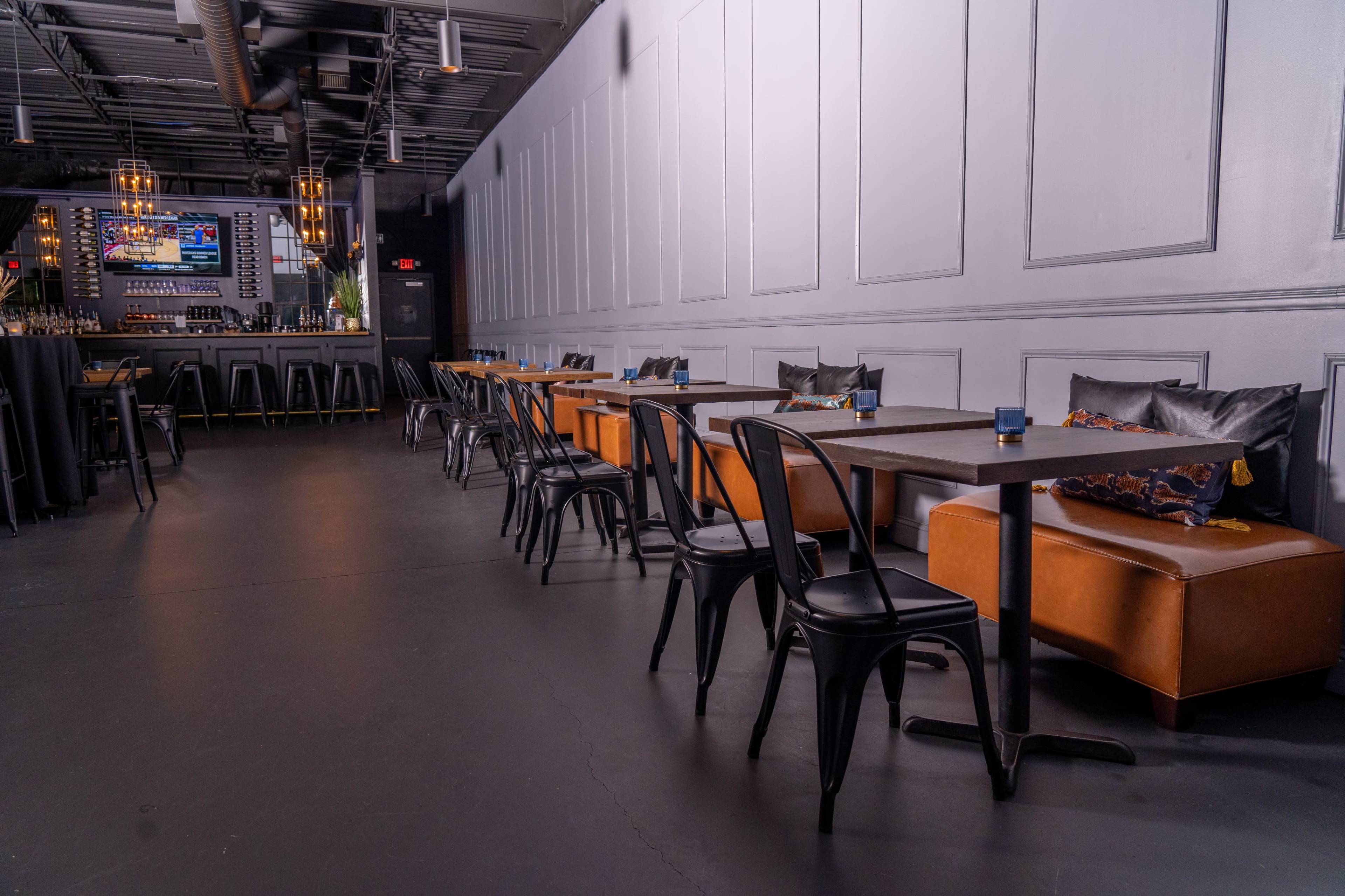 A modern restaurant interior with a row of brown cushioned benches and black metal chairs along sleek tables, leading to a bar area in the background.