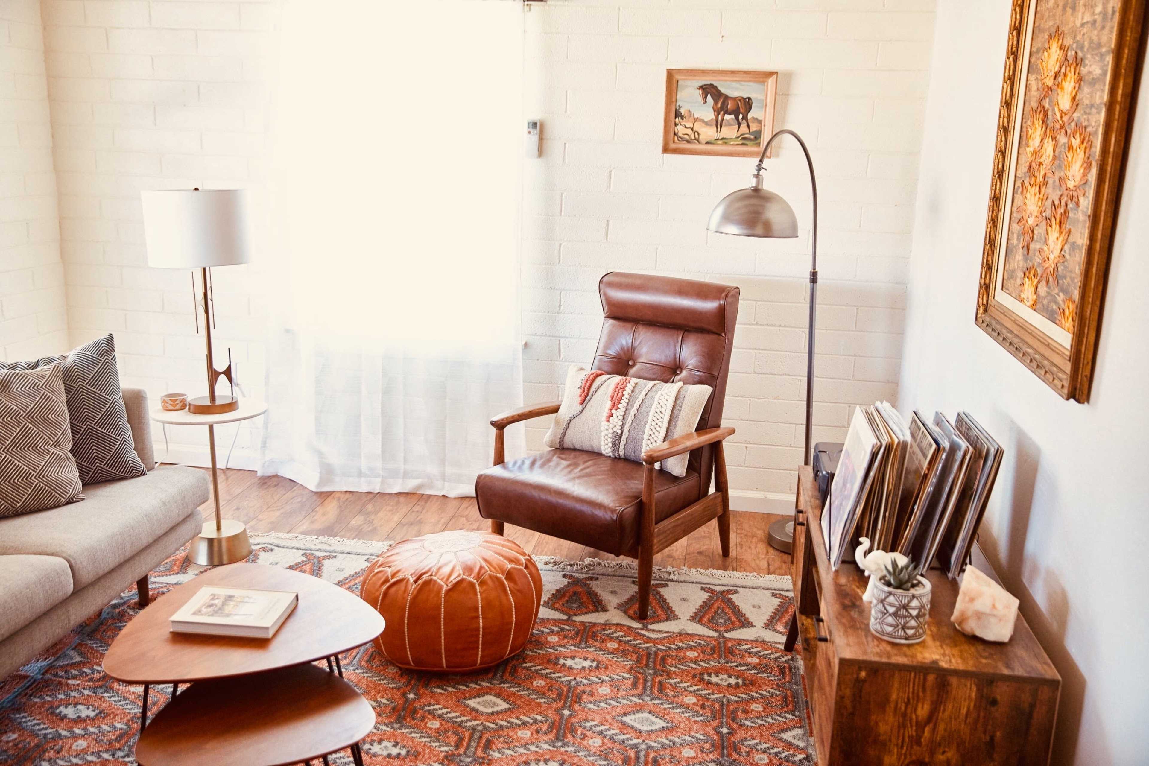 A spacious living room features a leather armchair, an ottoman, a side table, and a wooden storage unit filled with records, complemented by a large window and decorative wall art.