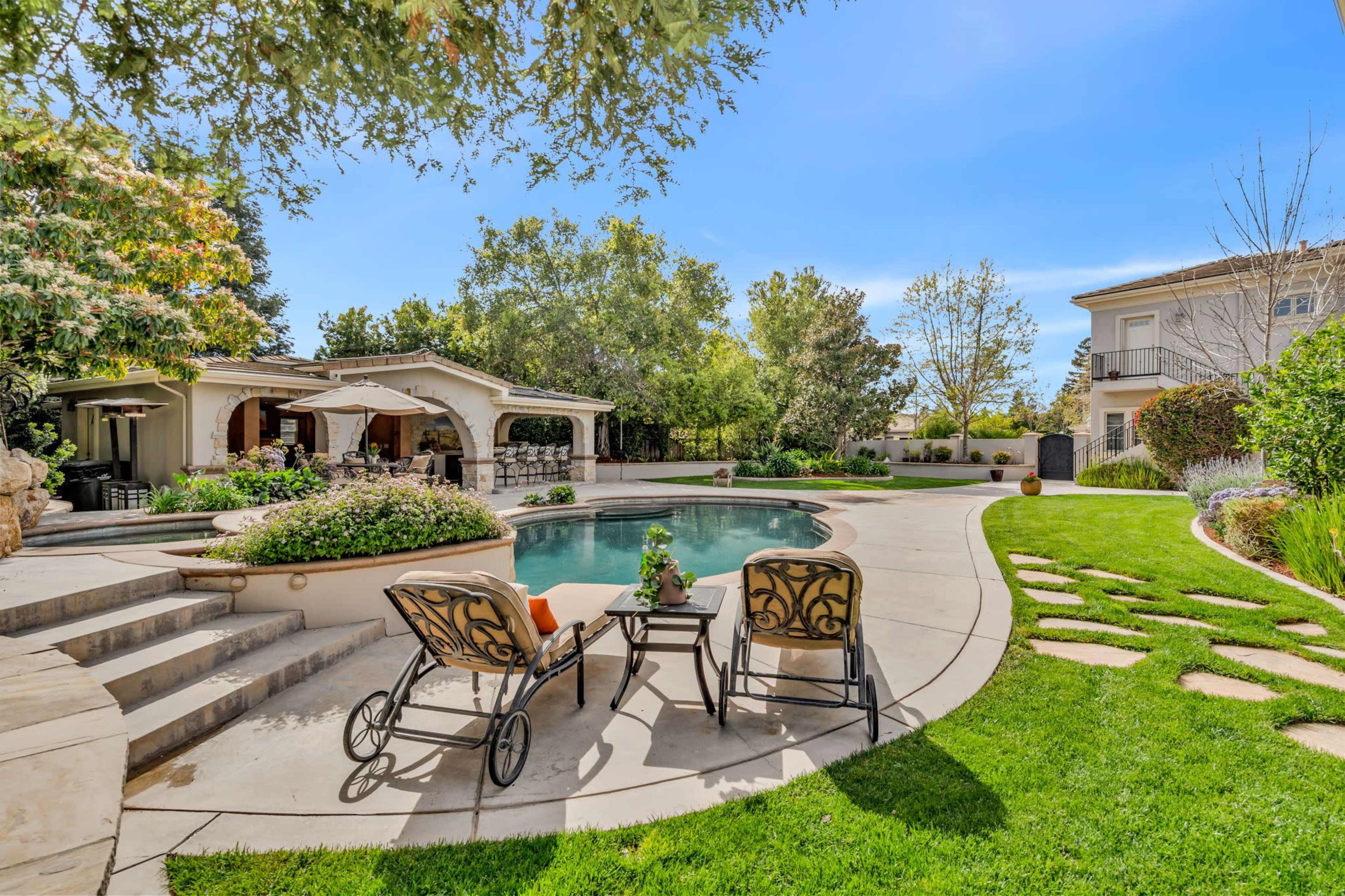 A backyard features a pool surrounded by landscaped gardens, stone steps, and seating areas with two chairs positioned near the water.