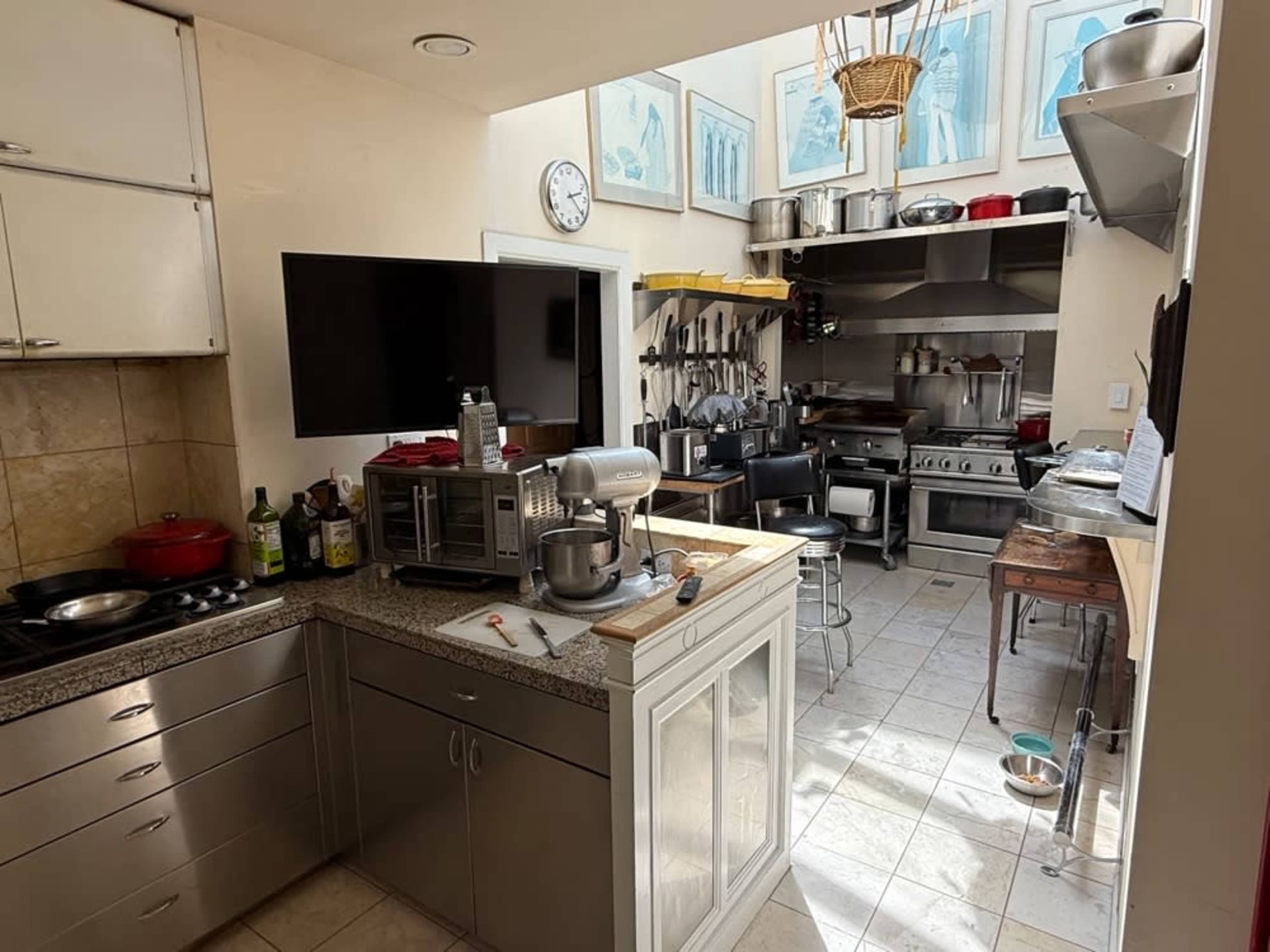The image shows a kitchen with stainless steel appliances, a central island with a mixer, and various cooking utensils hanging on the walls.