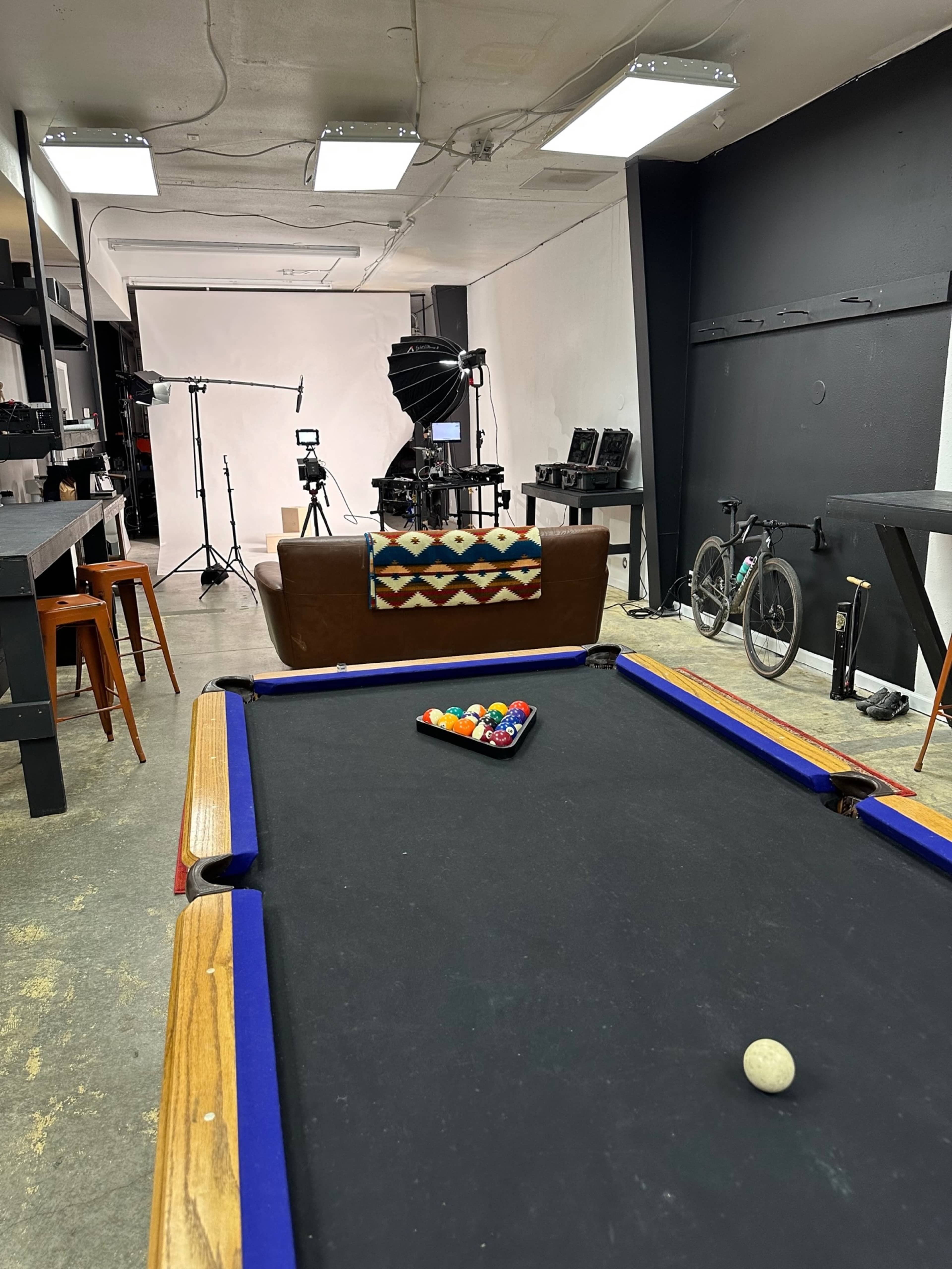 A room with a pool table in the foreground, a brown couch in the center, and various photography equipment and a bicycle in the background.