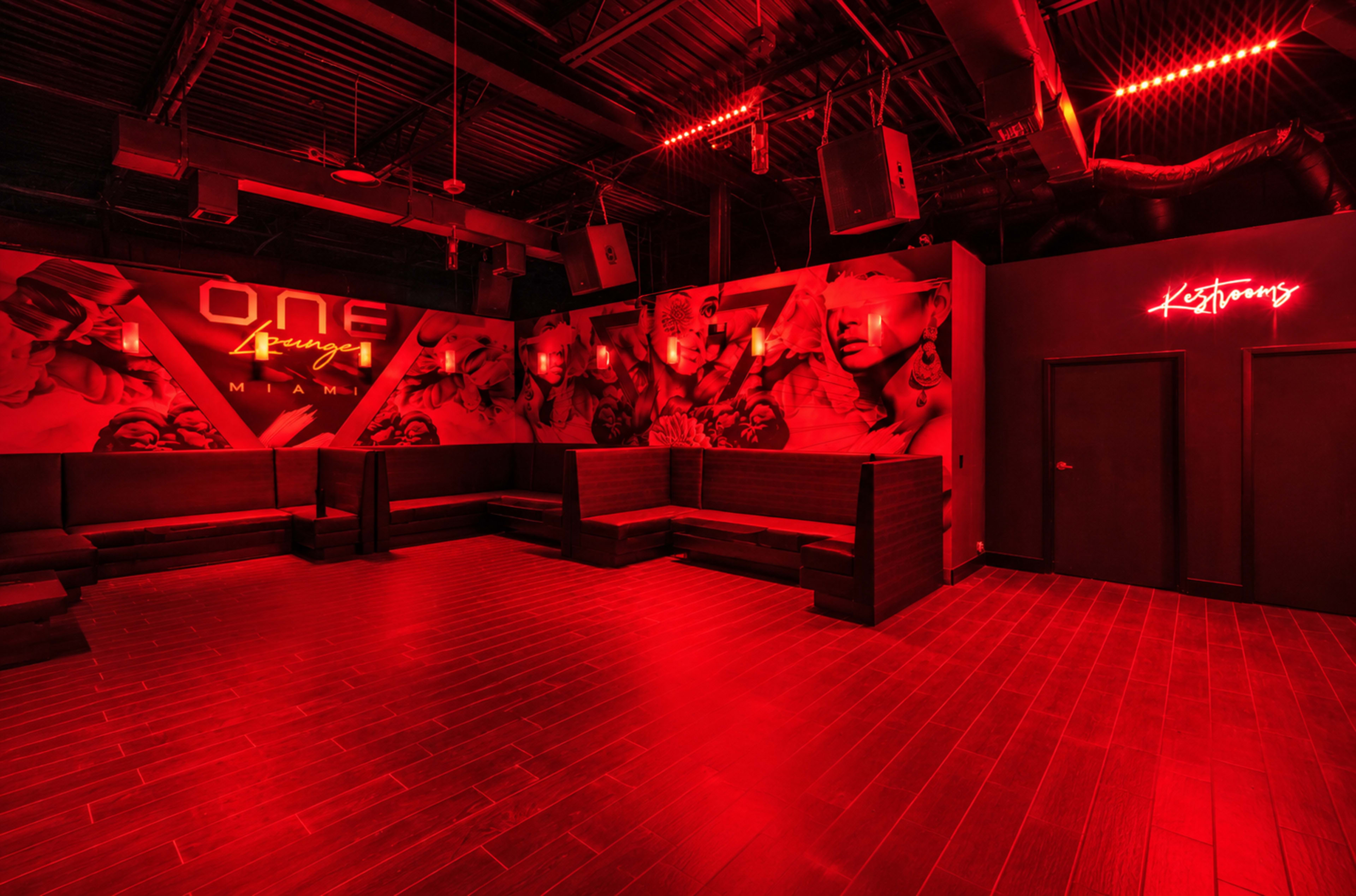 The image depicts a dimly lit venue with red lighting, featuring booths along the walls and a large mural of stylized faces.
