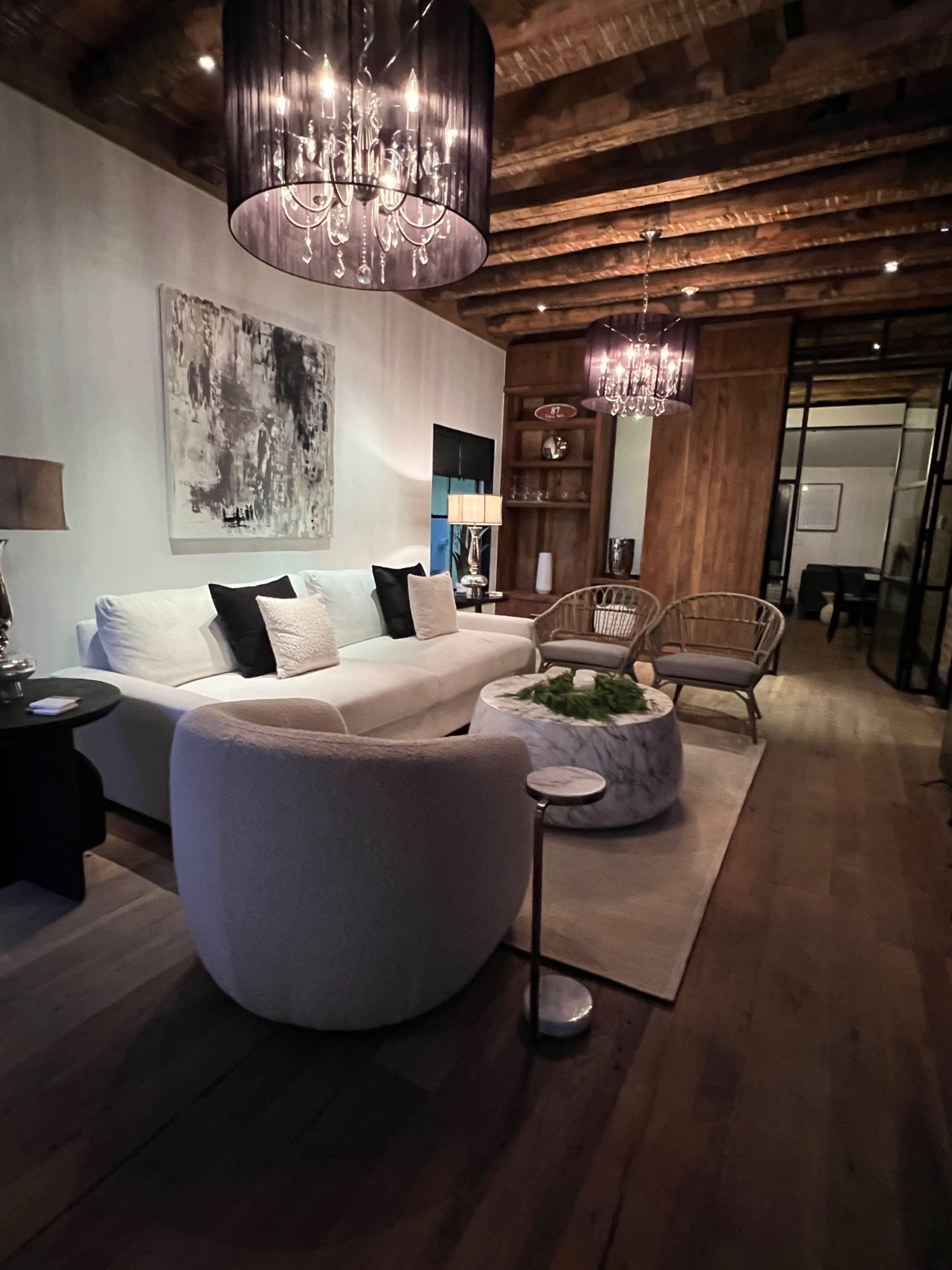 The image shows a living room with a white sofa, two wicker chairs, a marble coffee table, and chandeliers hanging from a wooden ceiling.