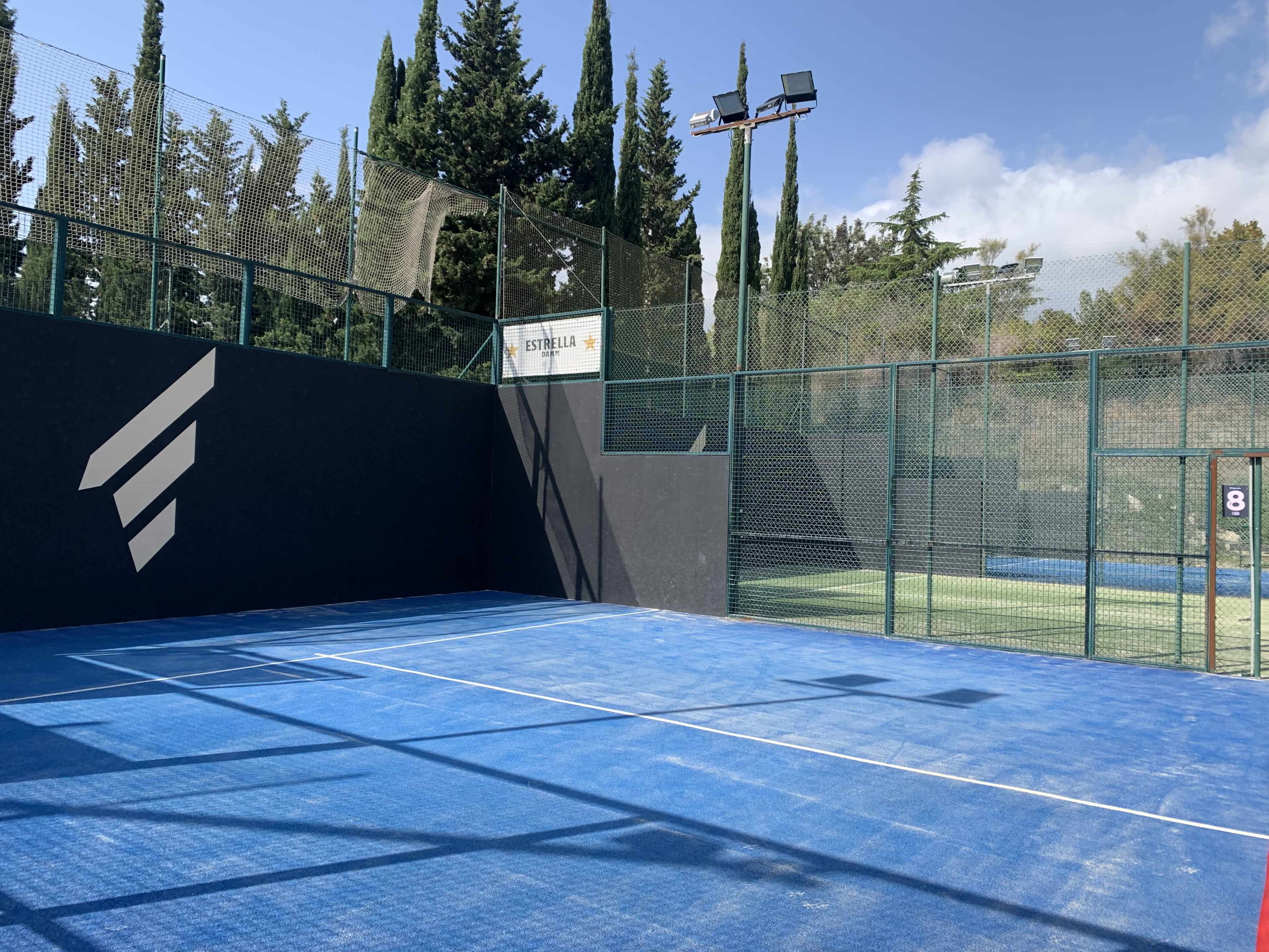 A blue padel court enclosed by a green mesh fence and surrounded by tall trees under a partly cloudy sky.