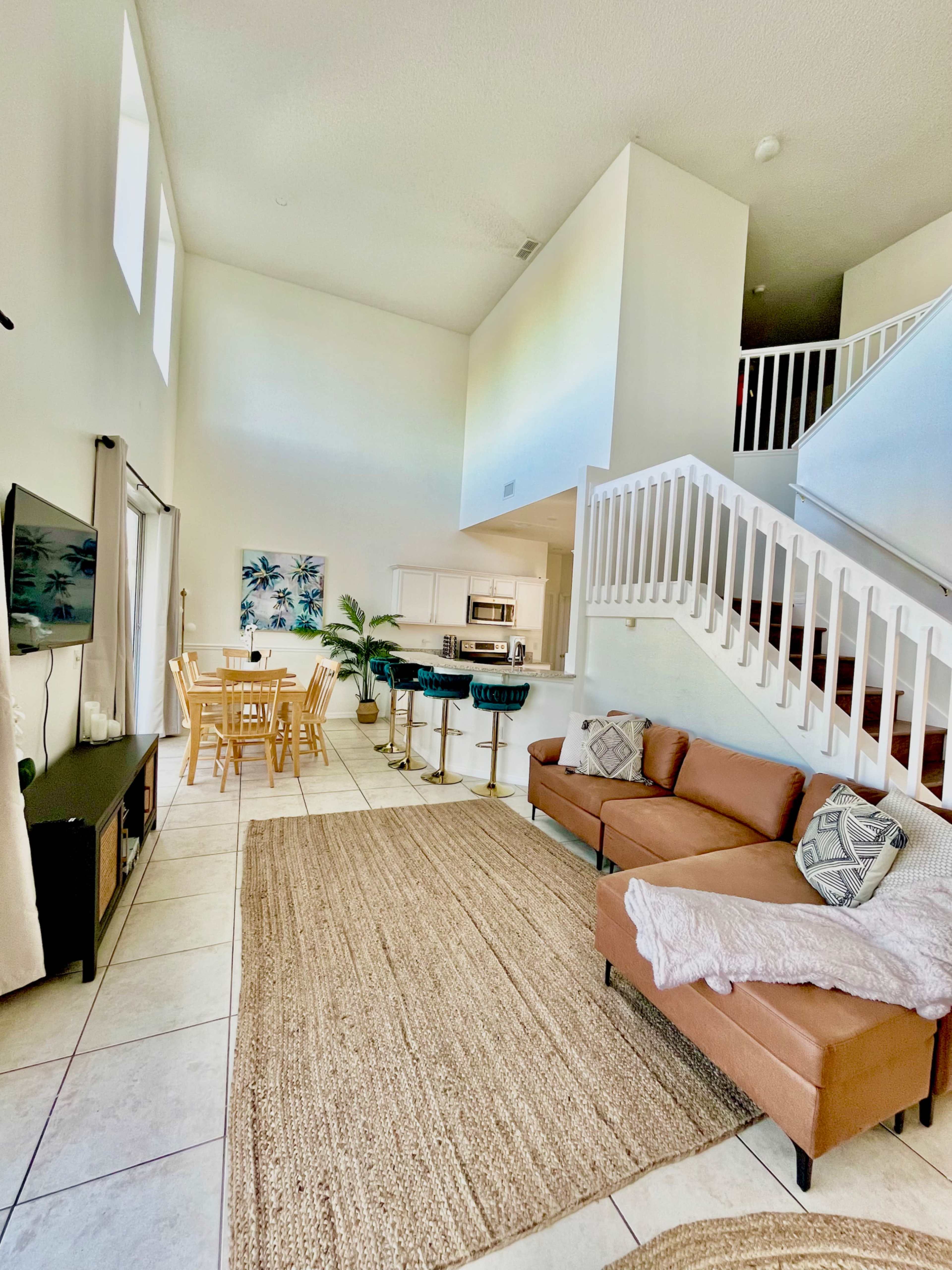 The image shows a brightly lit open living space with a staircase, a seating area with a brown couch, a dining table for six, and a kitchen visible in the background.